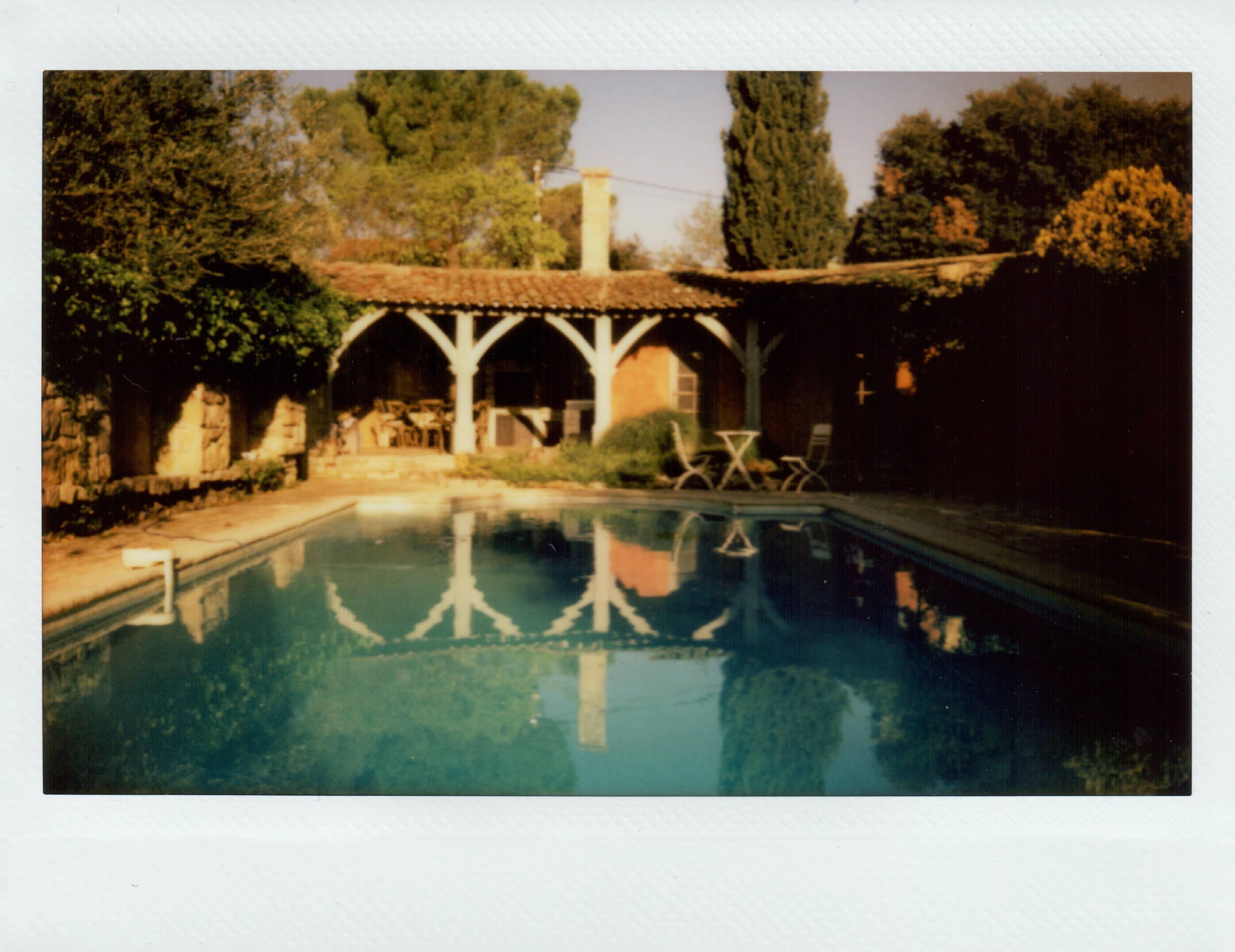 A swimming pool reflects a covered patio area surrounded by trees and shrubs.