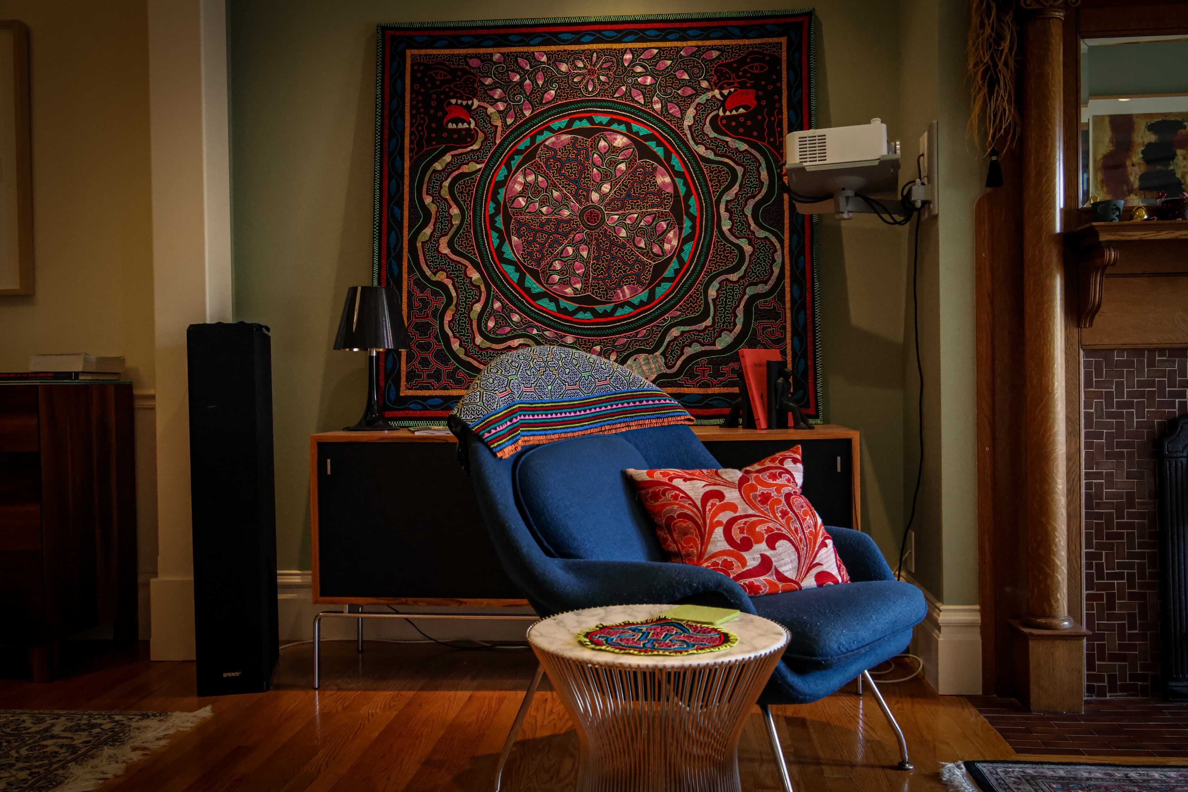 The scene features a blue chair with decorative pillows positioned in front of a colorful wall hanging and a media cabinet in a living room setting.