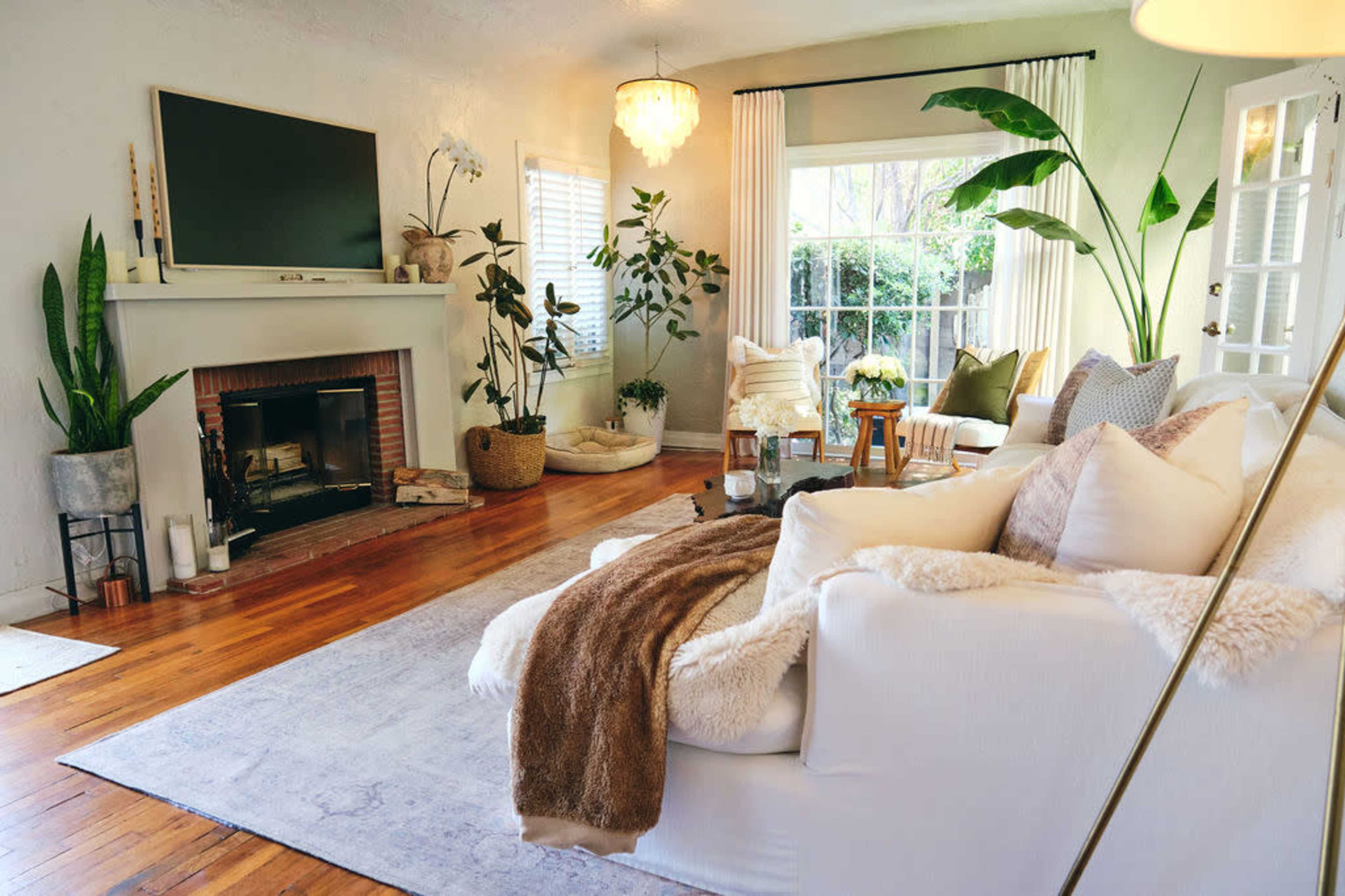 The image shows a cozy living room featuring a white sofa, a fireplace with a TV above it, potted plants, and large windows allowing natural light.
