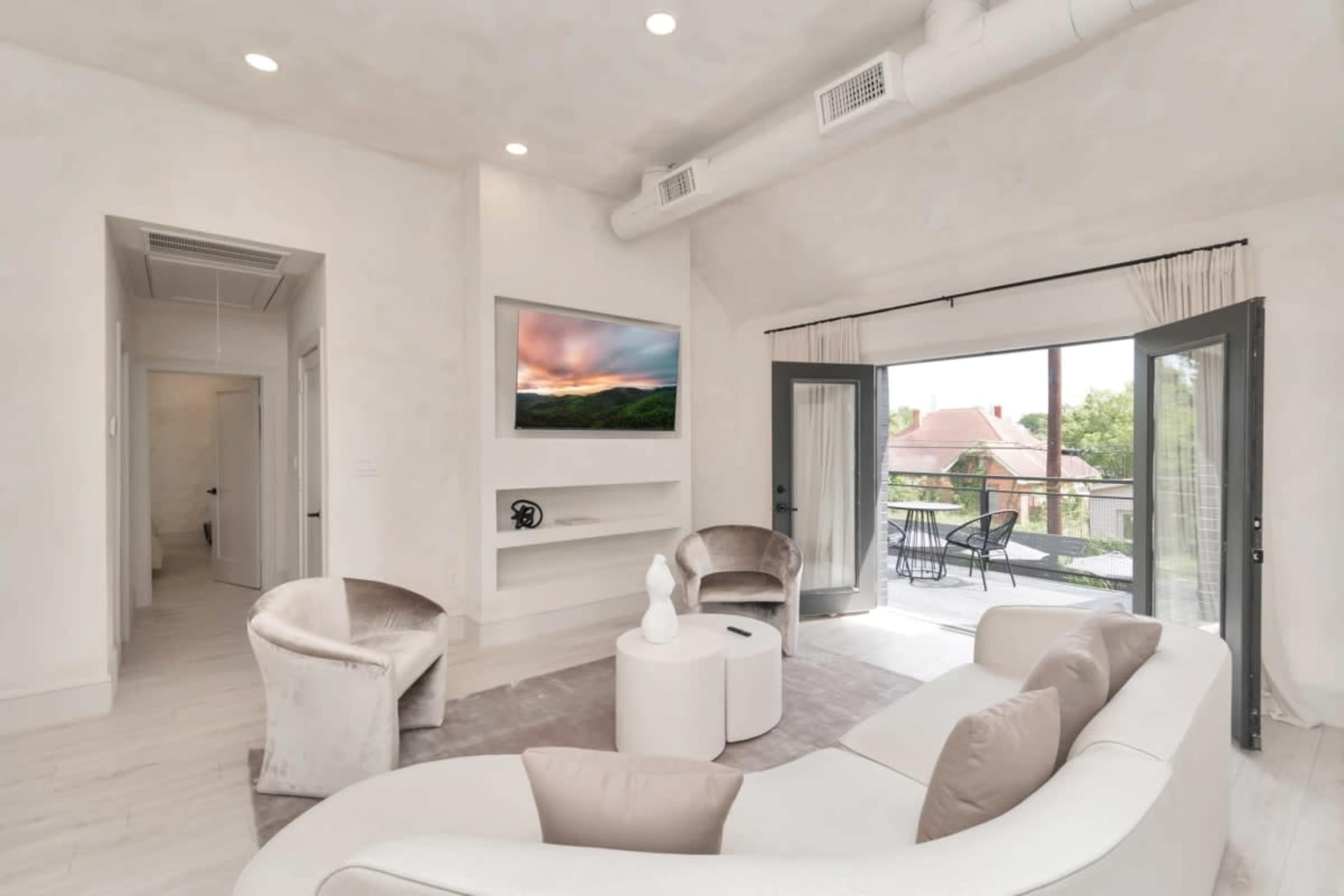 A modern living room with a curved sofa, two chairs, a coffee table, and a wall-mounted television, leading to a balcony through large glass doors.
