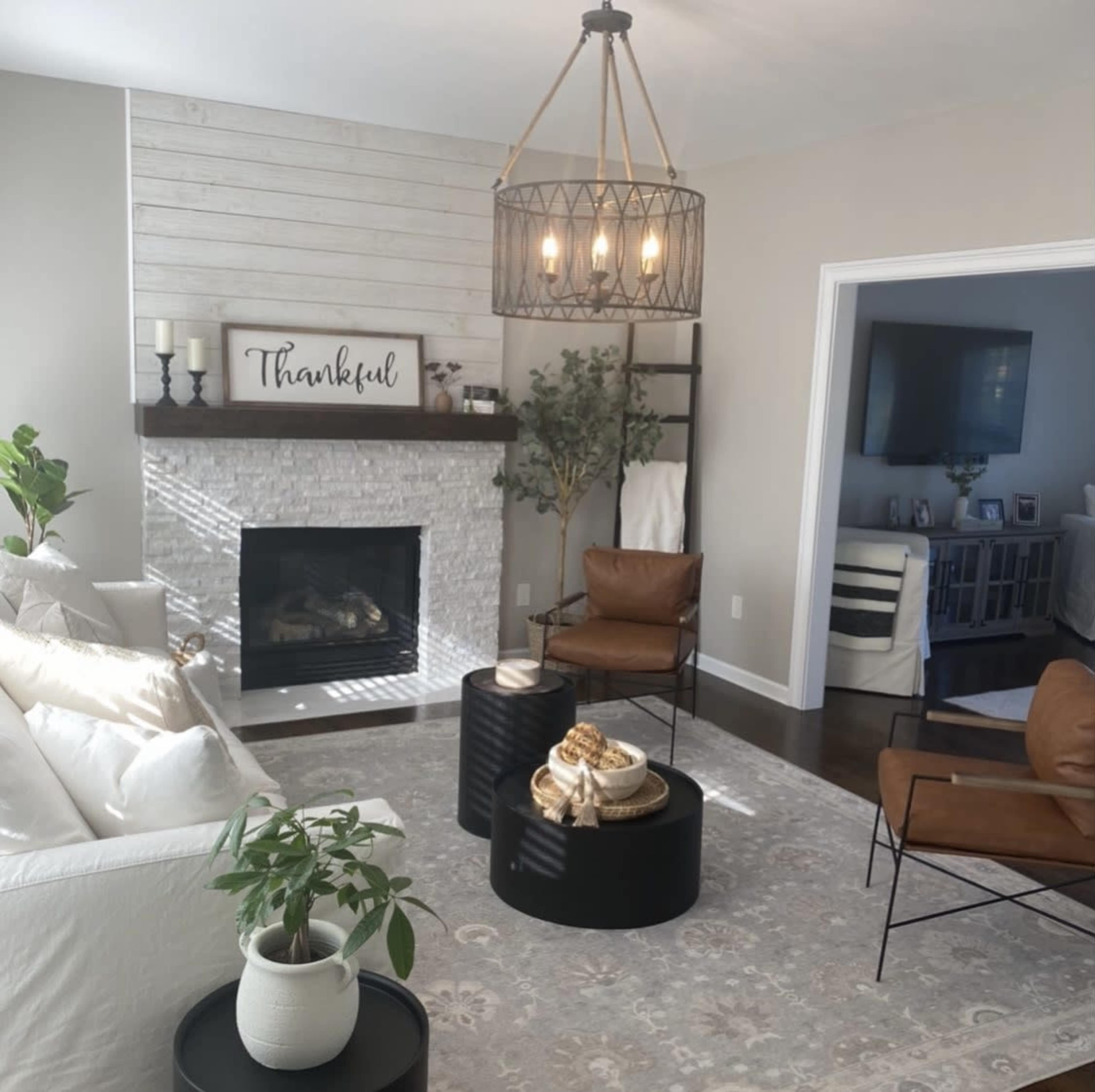 A cozy living room features a stone fireplace with a "Thankful" sign above, surrounded by a white sofa, brown chairs, and a decorative rug.