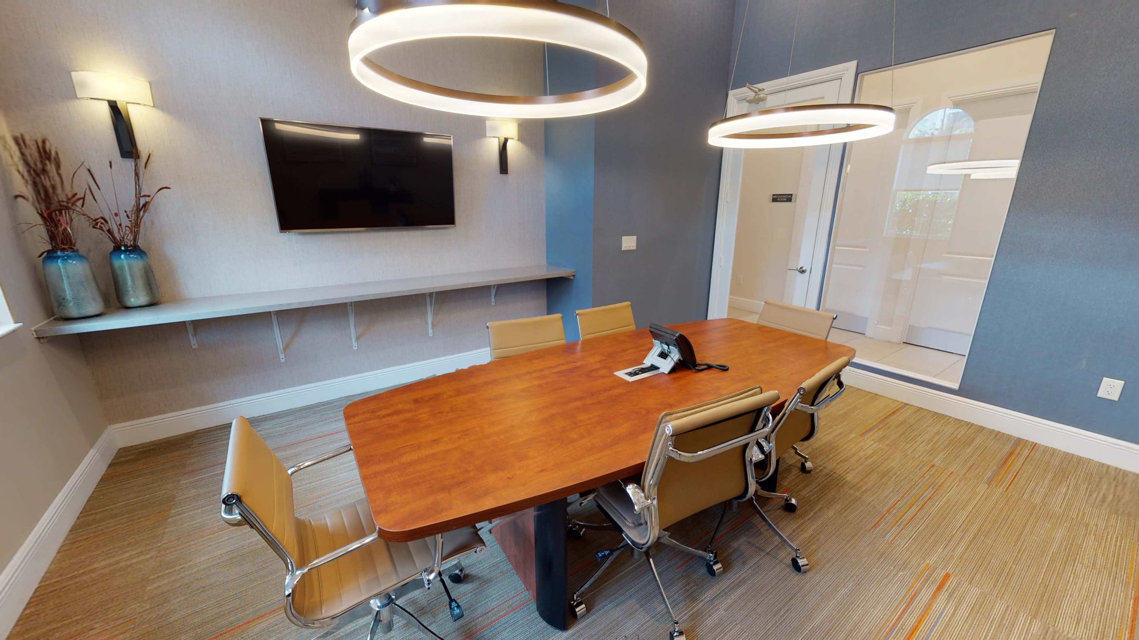 A modern conference room featuring a wooden table, six chairs, a wall-mounted television, and two pendant lights.