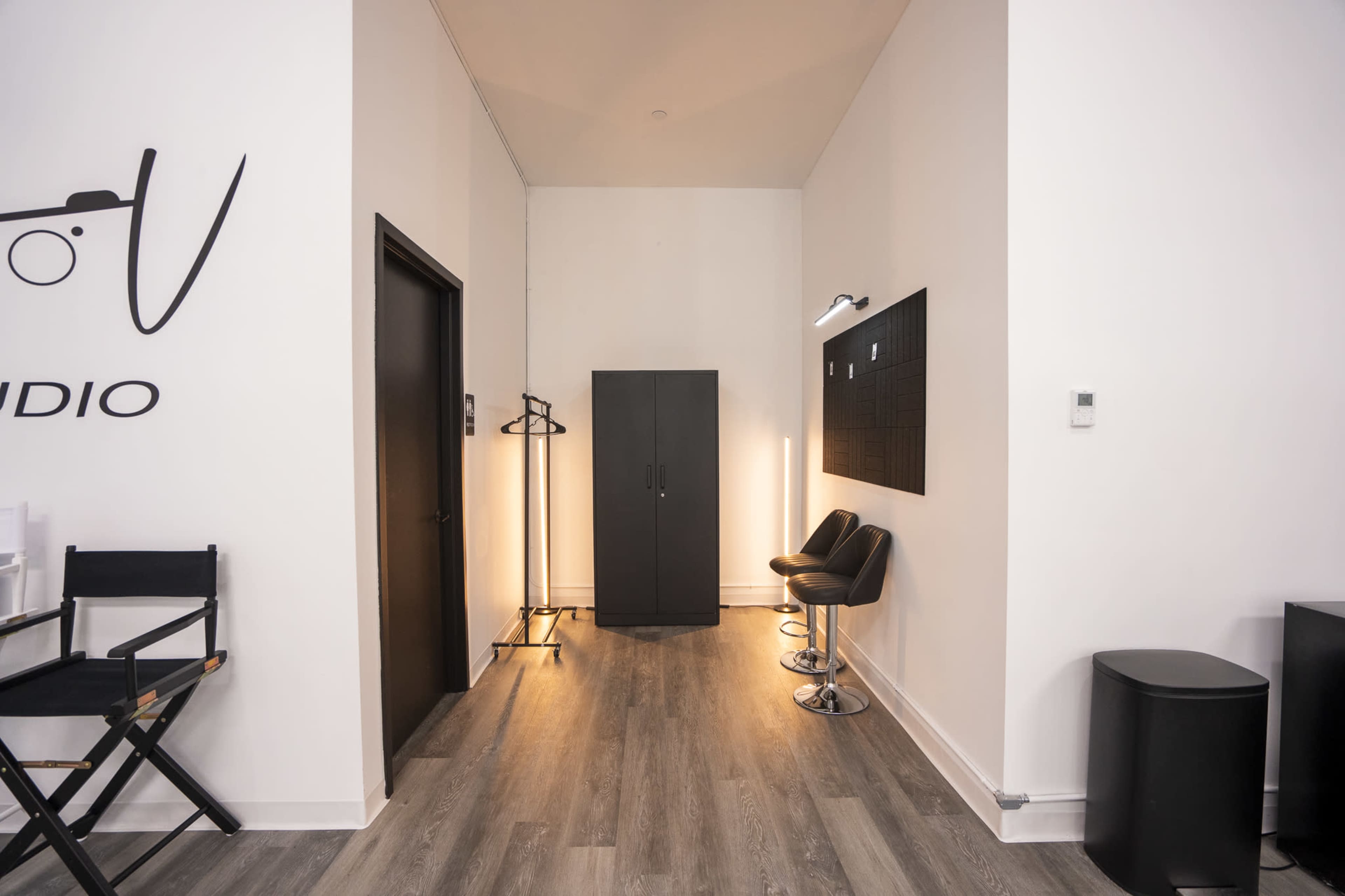 The image shows a minimalist studio interior with a black wardrobe, two barstools, and a director's chair against a white wall.