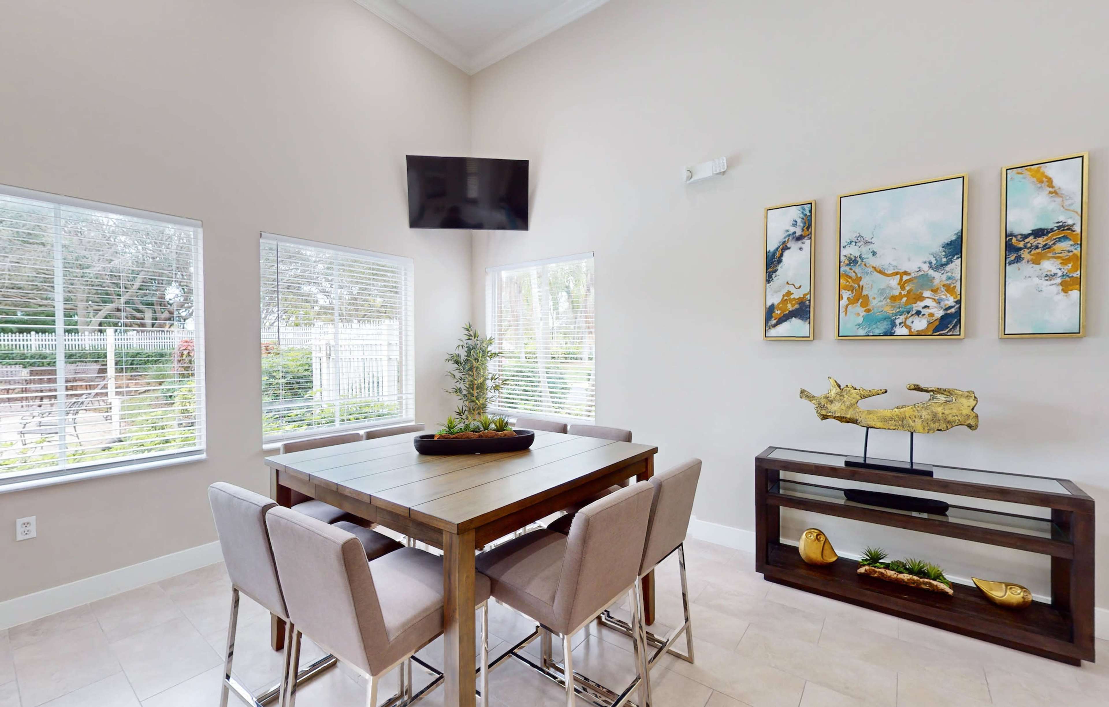 A dining area features a wooden table with four upholstered chairs, a wall-mounted TV, and decorative artwork above a console table.