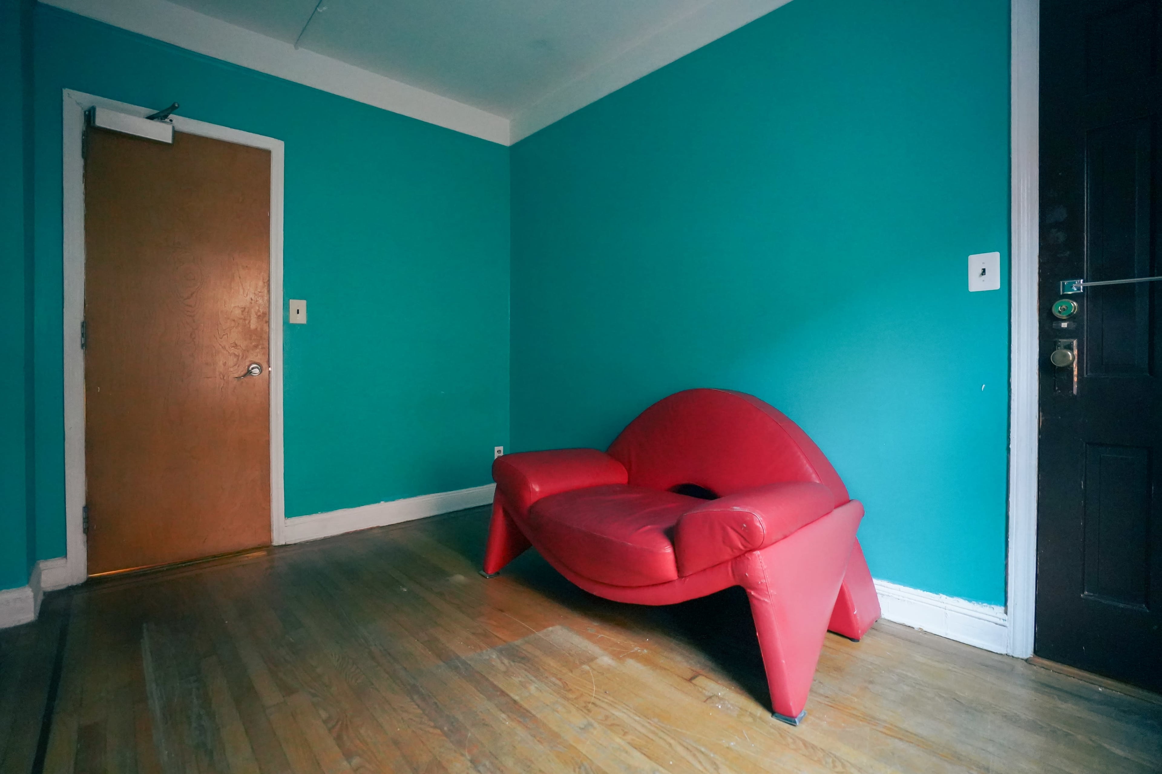 The image shows a sparsely furnished room with a bright turquoise wall, a wooden door, and a red, uniquely shaped sofa.