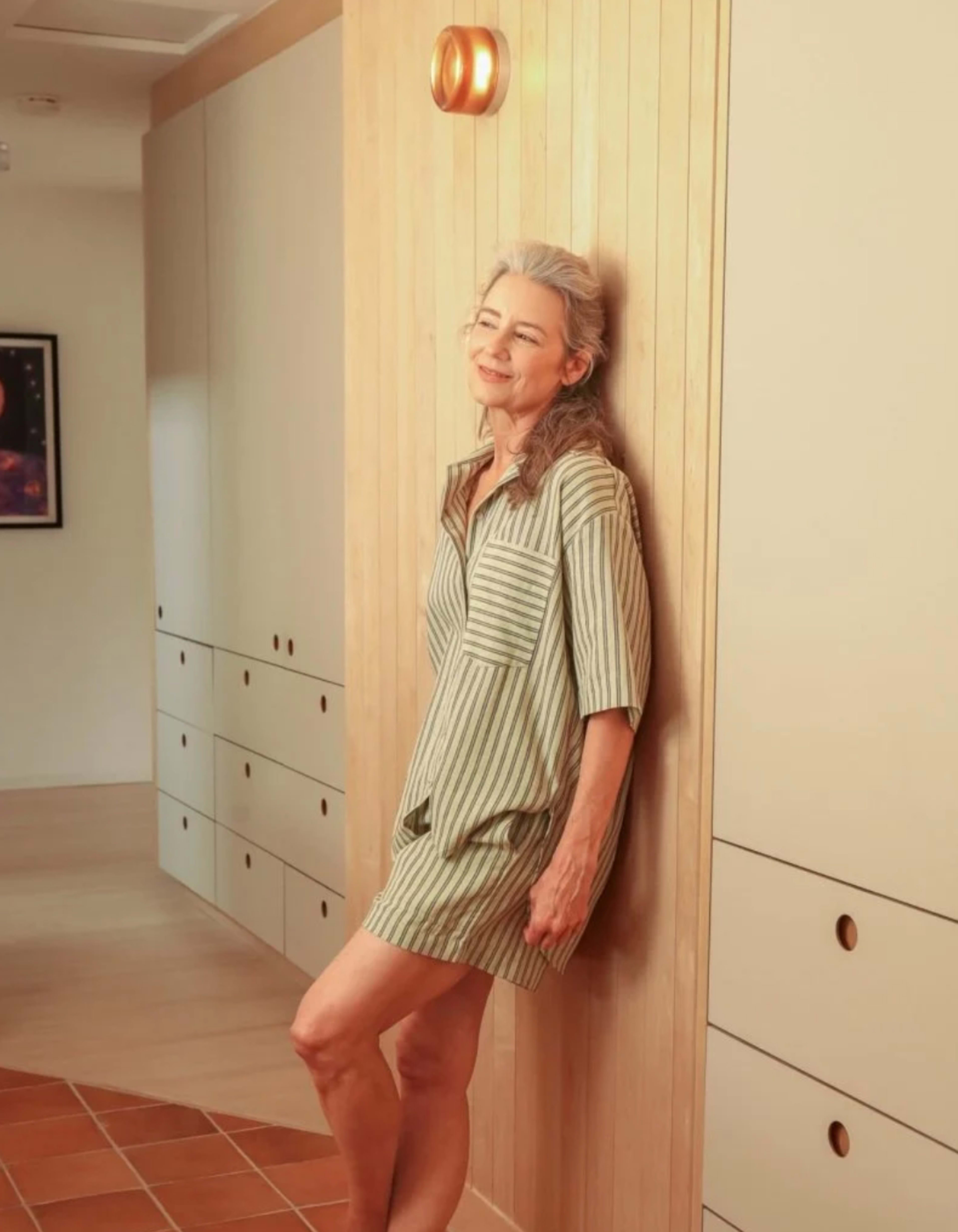 A woman with long gray hair stands against a wooden wall in a light-colored room with built-in cabinets.