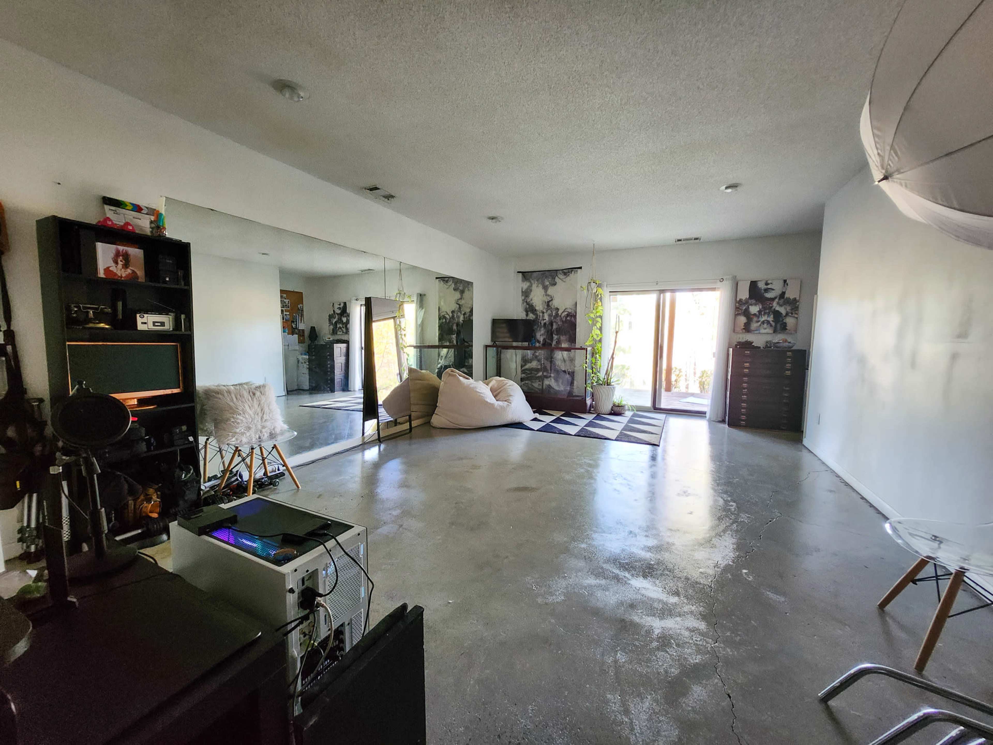 The image shows a spacious living room with a large mirror, a gray rug, beanbag chairs, and a view of a patio through sliding glass doors.