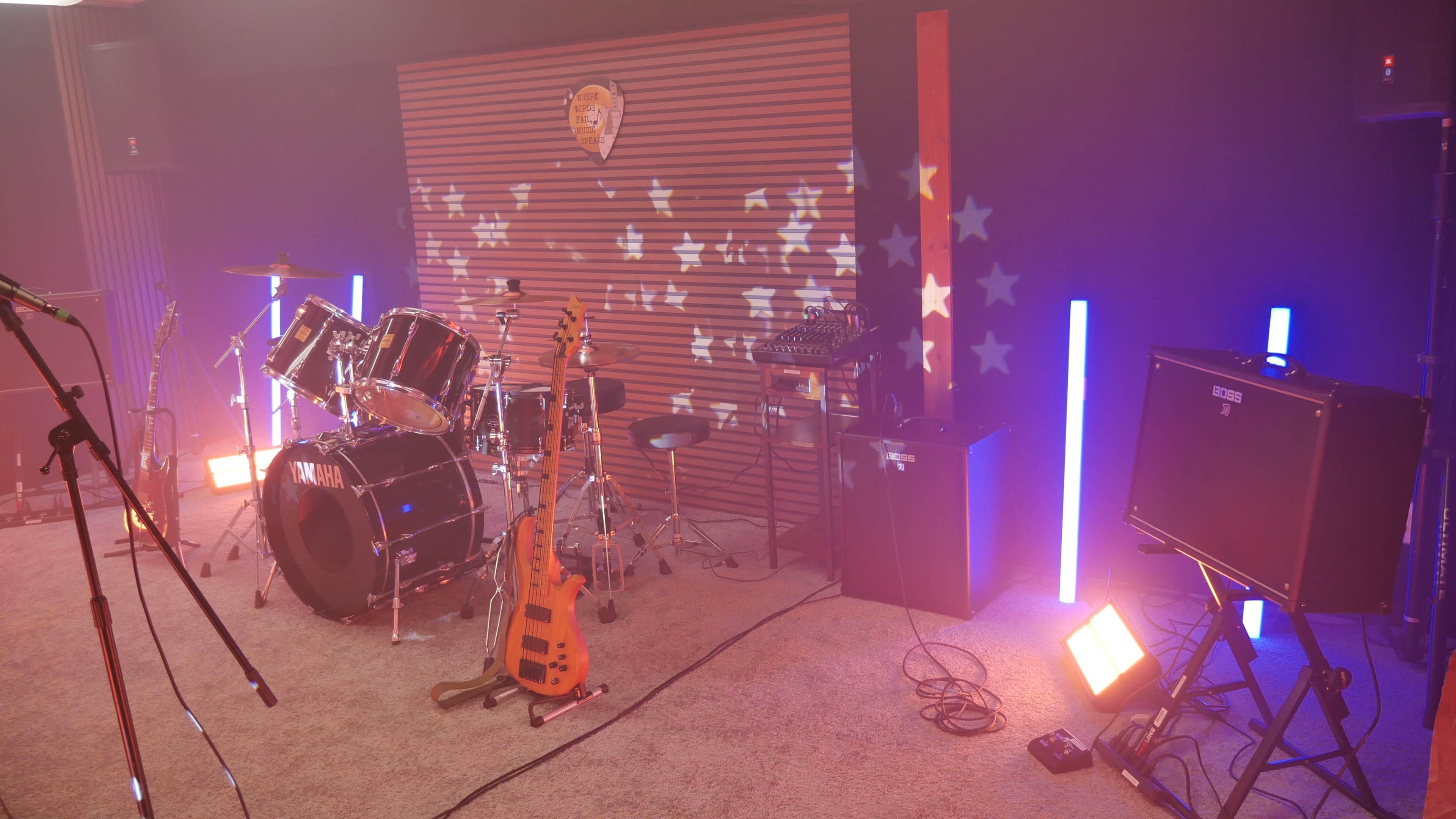 The scene shows a music rehearsal space featuring a drum set, electric guitar, amplifiers, and colorful stage lighting.