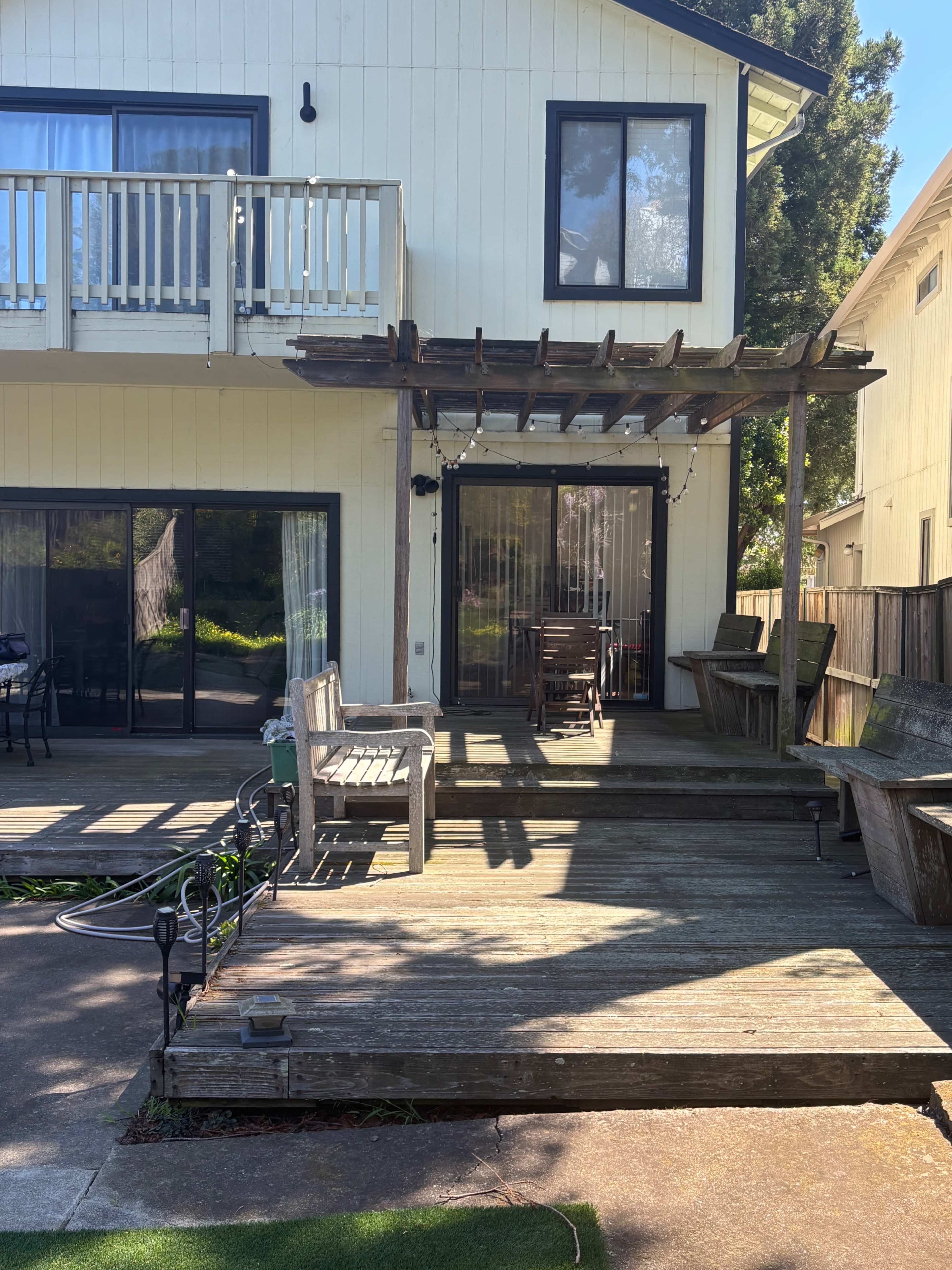 The image shows the back exterior of a two-story house with a balcony, a wooden deck, and a pergola over a seating area.