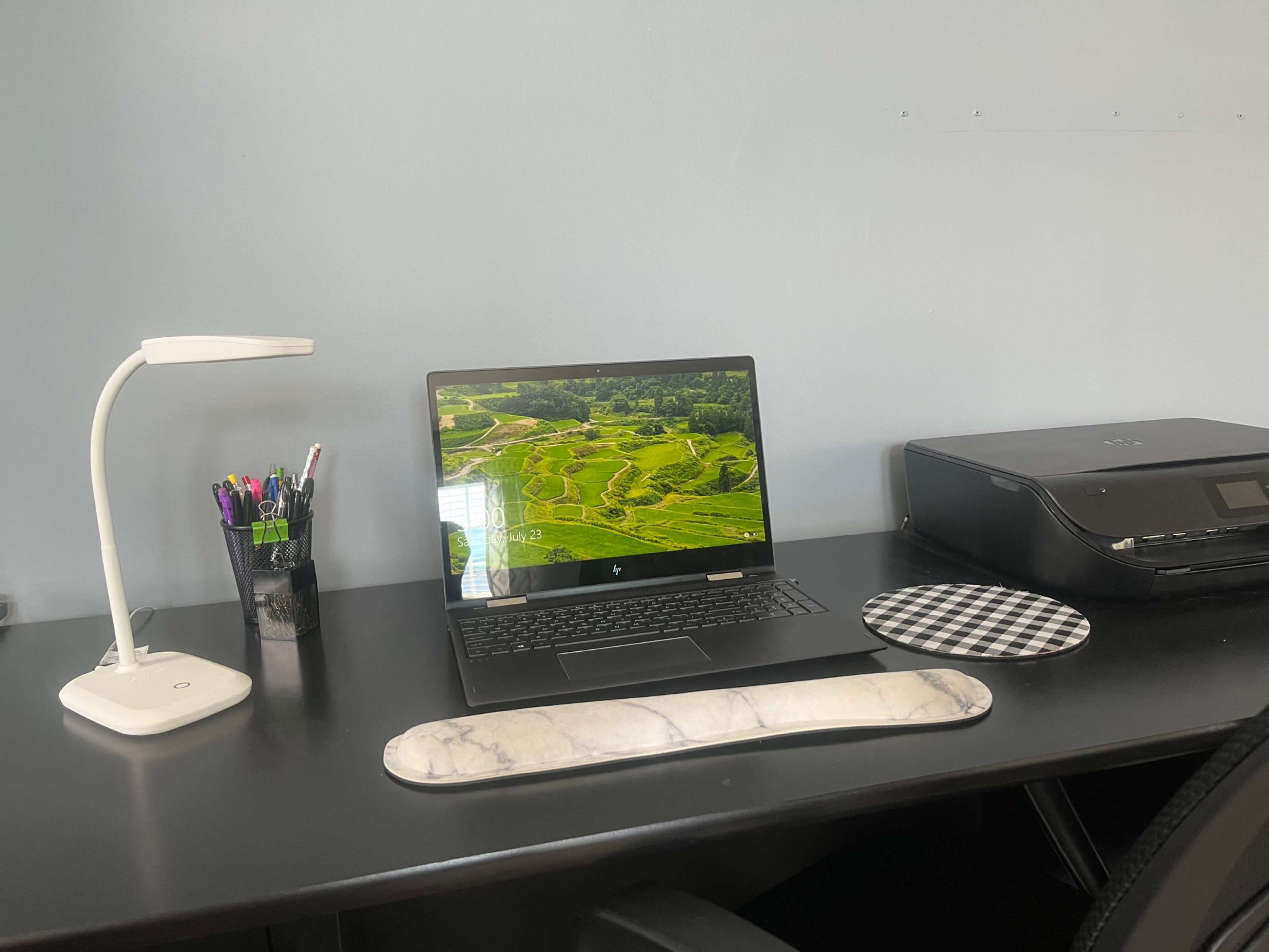 A laptop displaying a landscape image is placed on a black desk alongside a lamp, a printer, and various writing utensils in a cup.