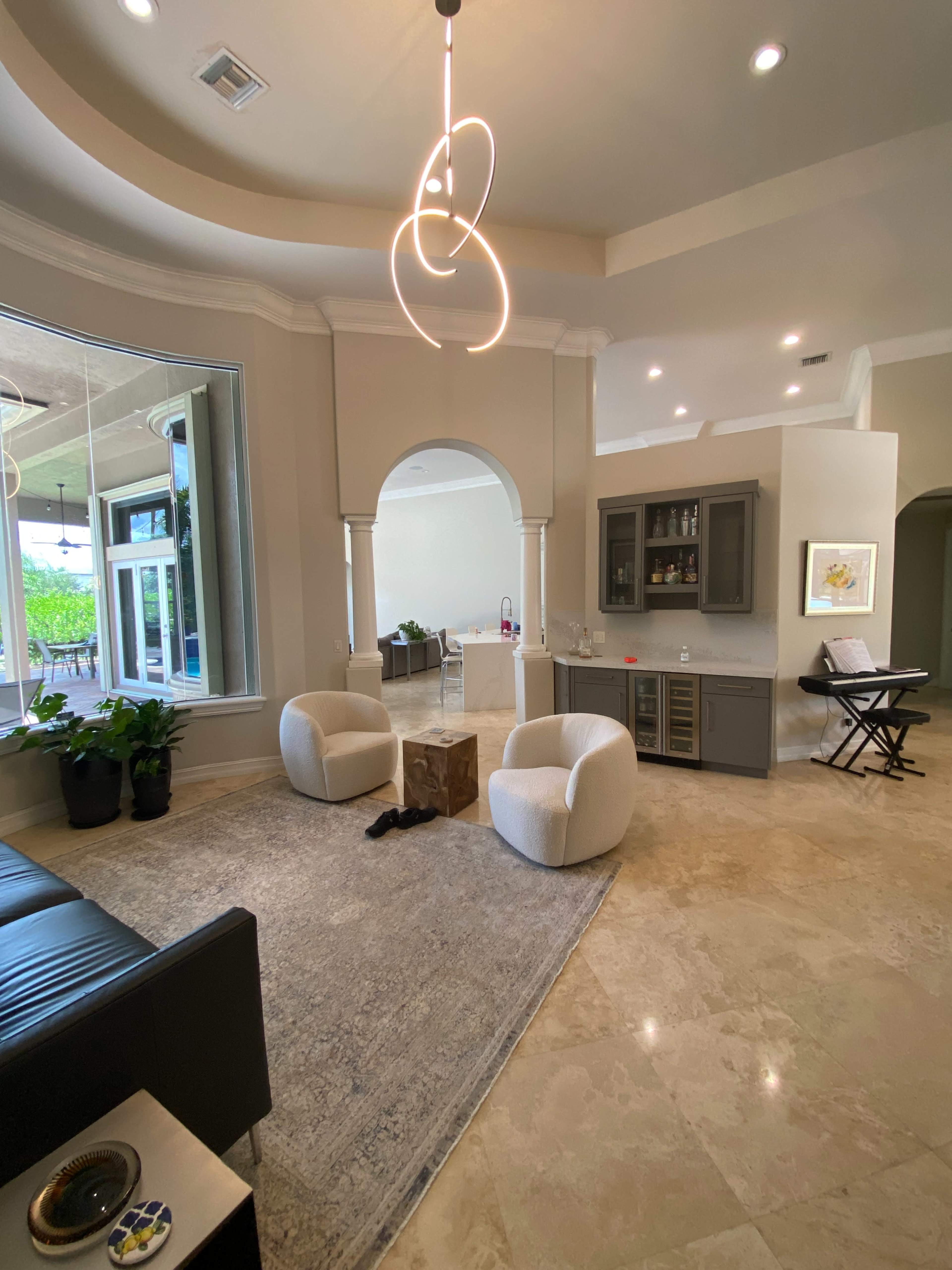 A spacious living area featuring two round armchairs, a small wooden table, and a bar area with cabinetry, all set on a light-colored stone floor.