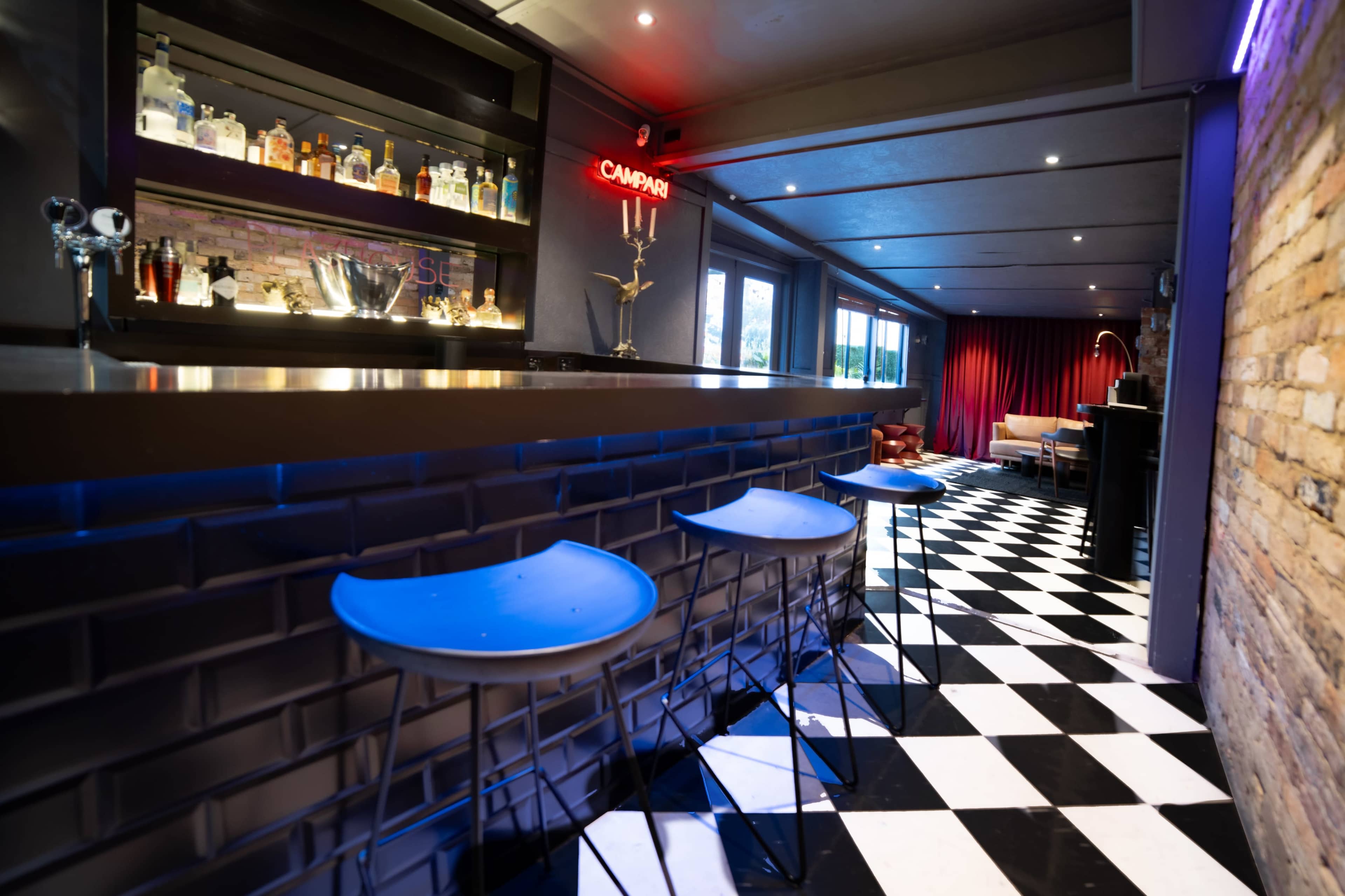 The image depicts a stylish bar area with a black and white checkered floor, illuminated shelves displaying various bottles, and three blue bar stools in front of a sleek counter.