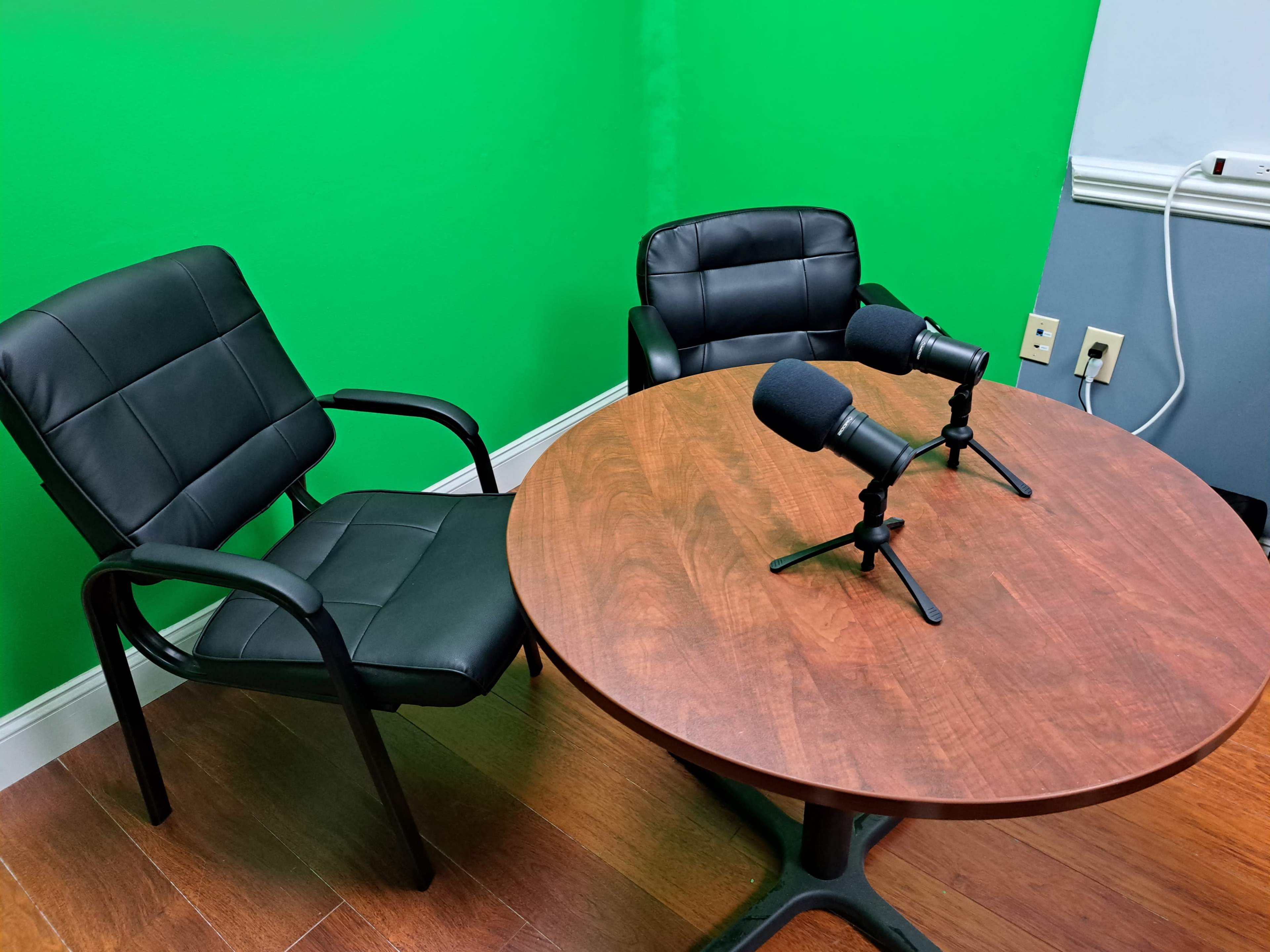 Two black chairs positioned around a circular wooden table with two microphones on stands, set against a green backdrop.
