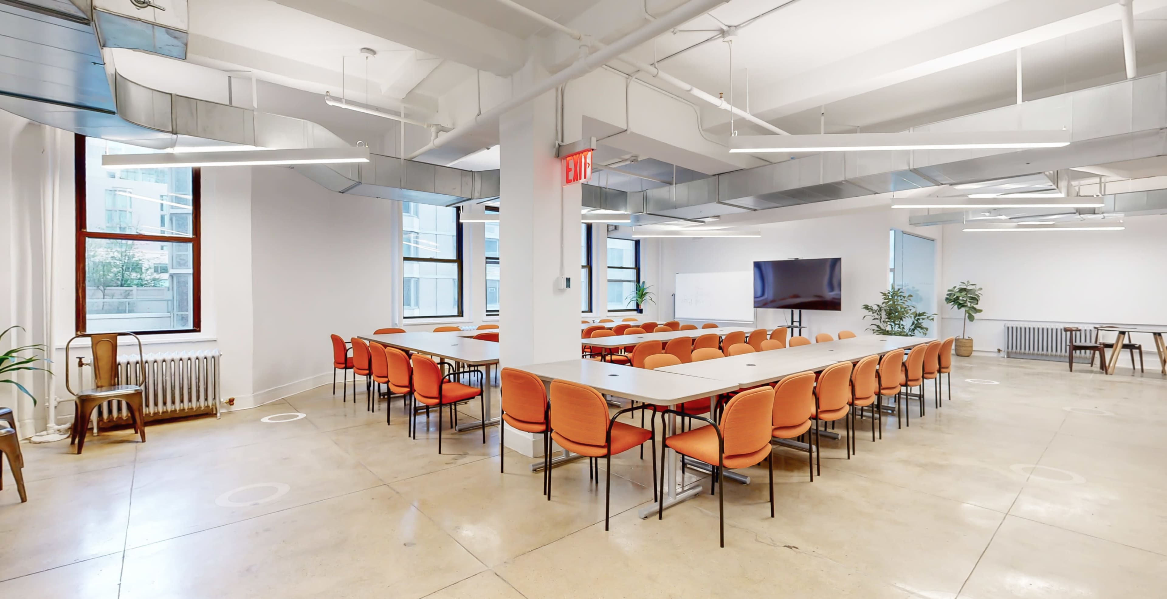 A modern conference room with rows of orange chairs arranged around long tables, large windows providing natural light, and a television screen on one wall.