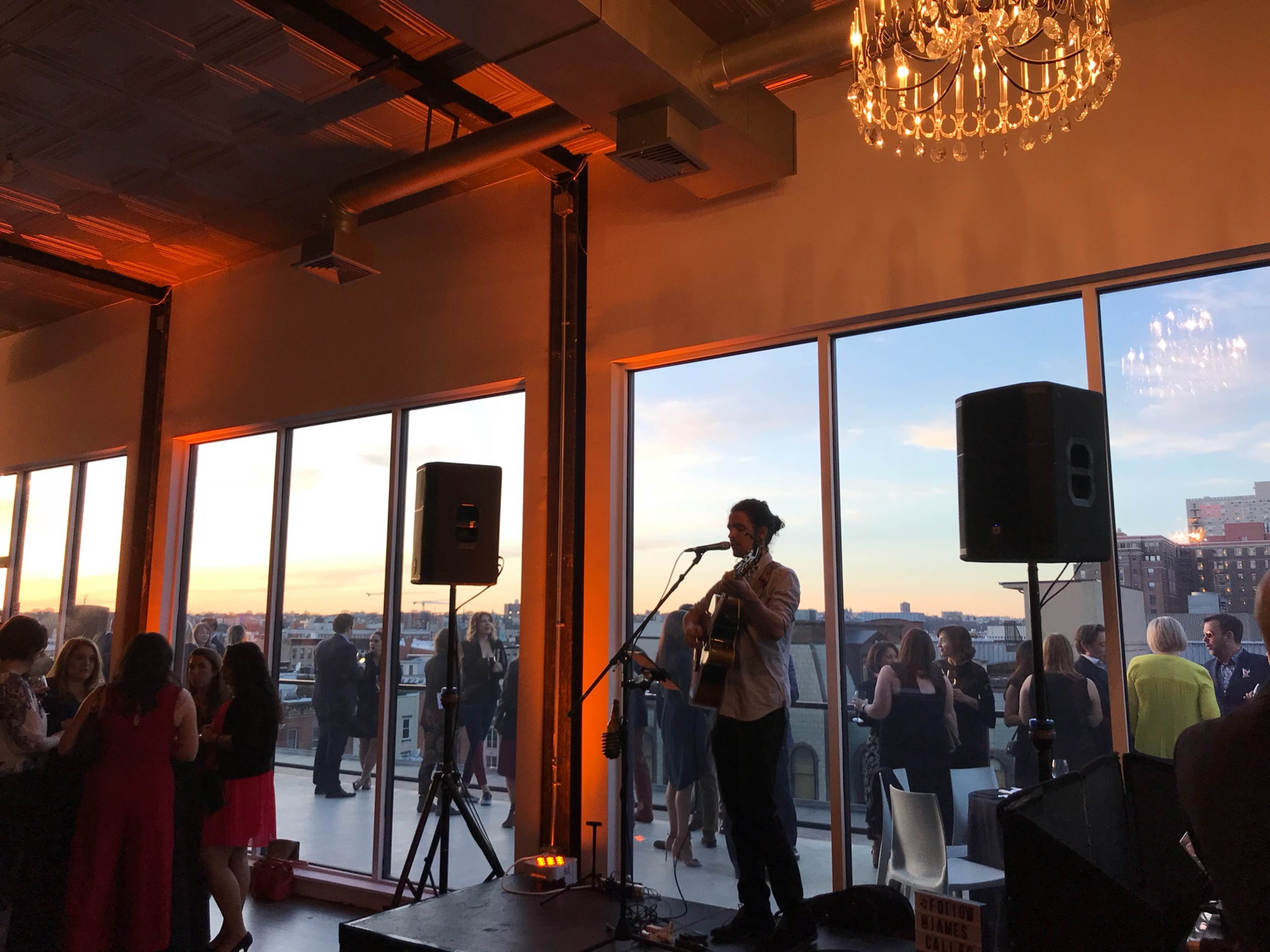 A musician plays guitar on a stage in front of large windows, overlooking a city skyline at sunset, while guests mingle at a reception.
