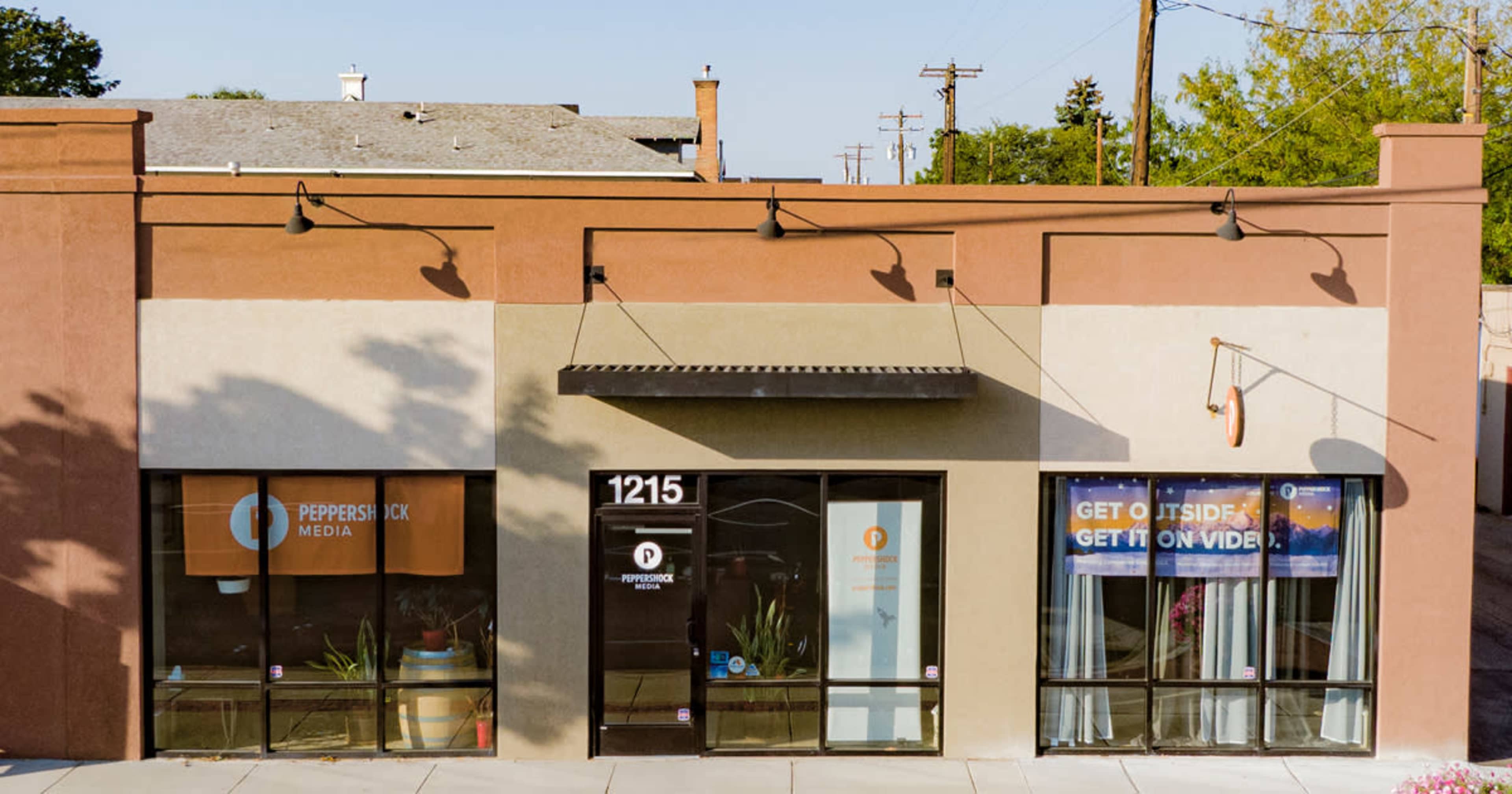 A one-story commercial building with large windows, featuring signage for "Pepperstick Media" and a promotional message urging visitors to "GET OUTSIDE, GET ON VIDEO."