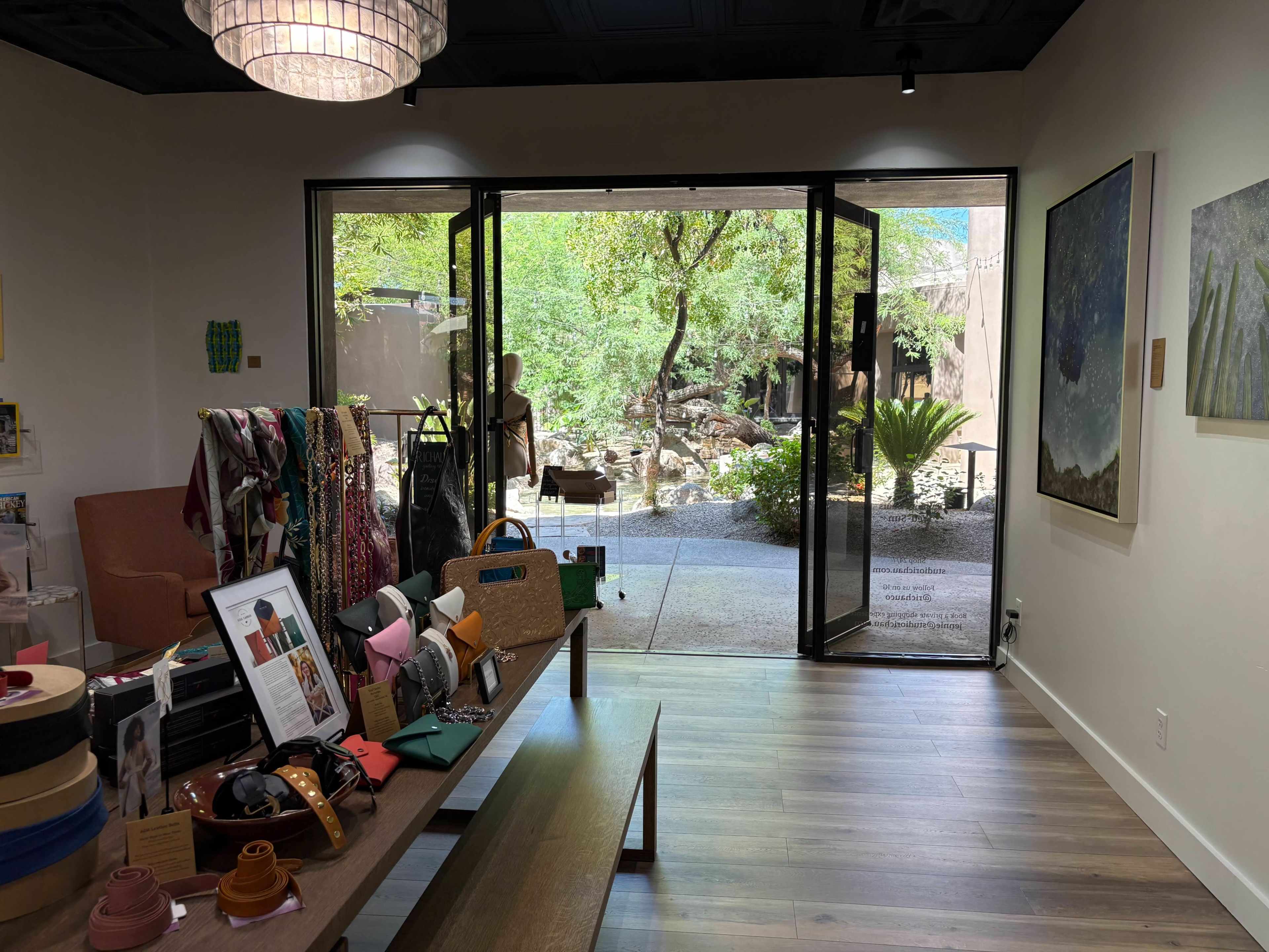 The interior of a retail space features a display of various accessories and clothing items, with large glass doors opening to a scenic outdoor area that includes greenery and a water feature.