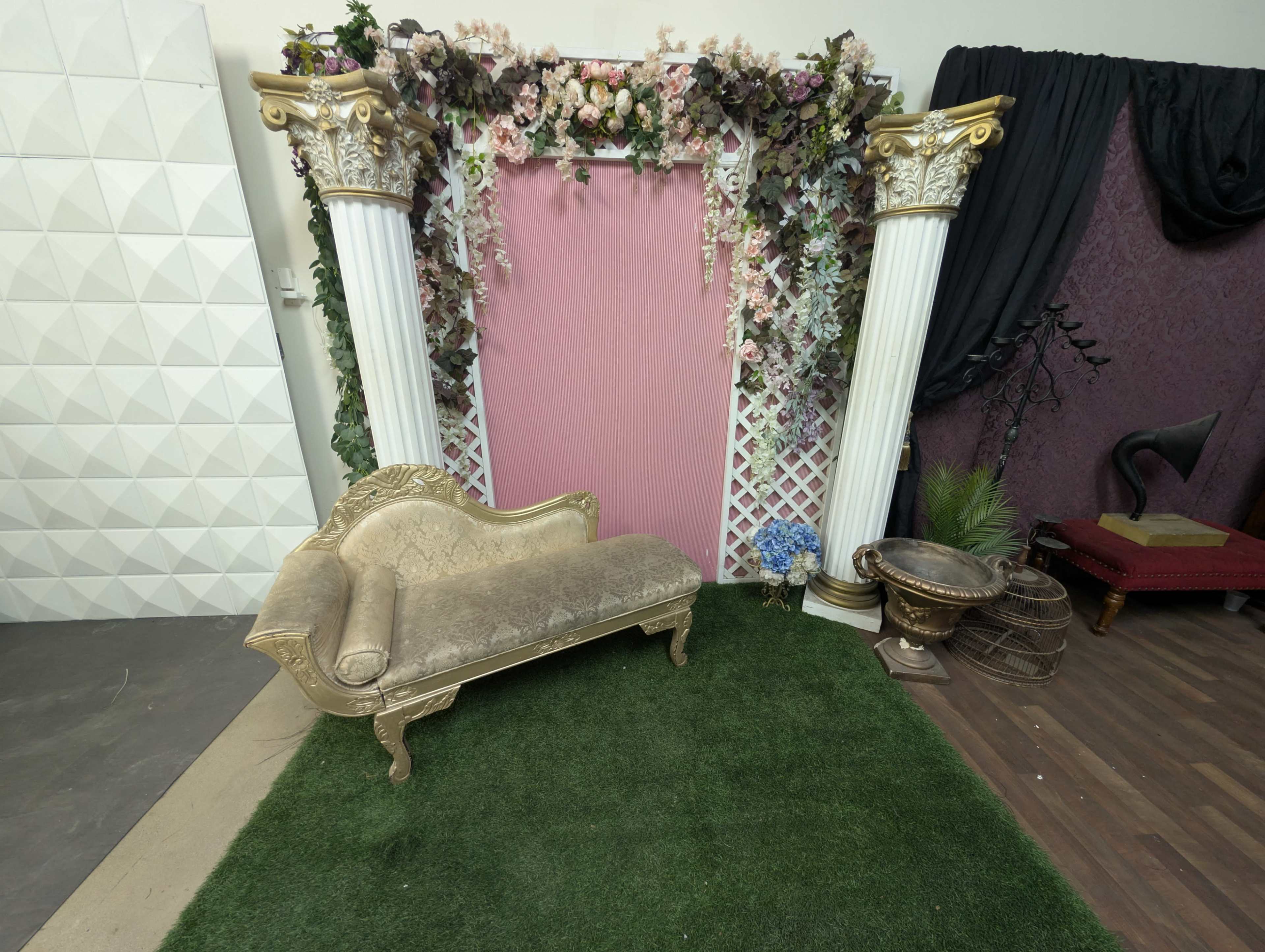 The scene features an ornate backdrop with two tall columns draped in flowers and a settee on a patch of artificial grass.