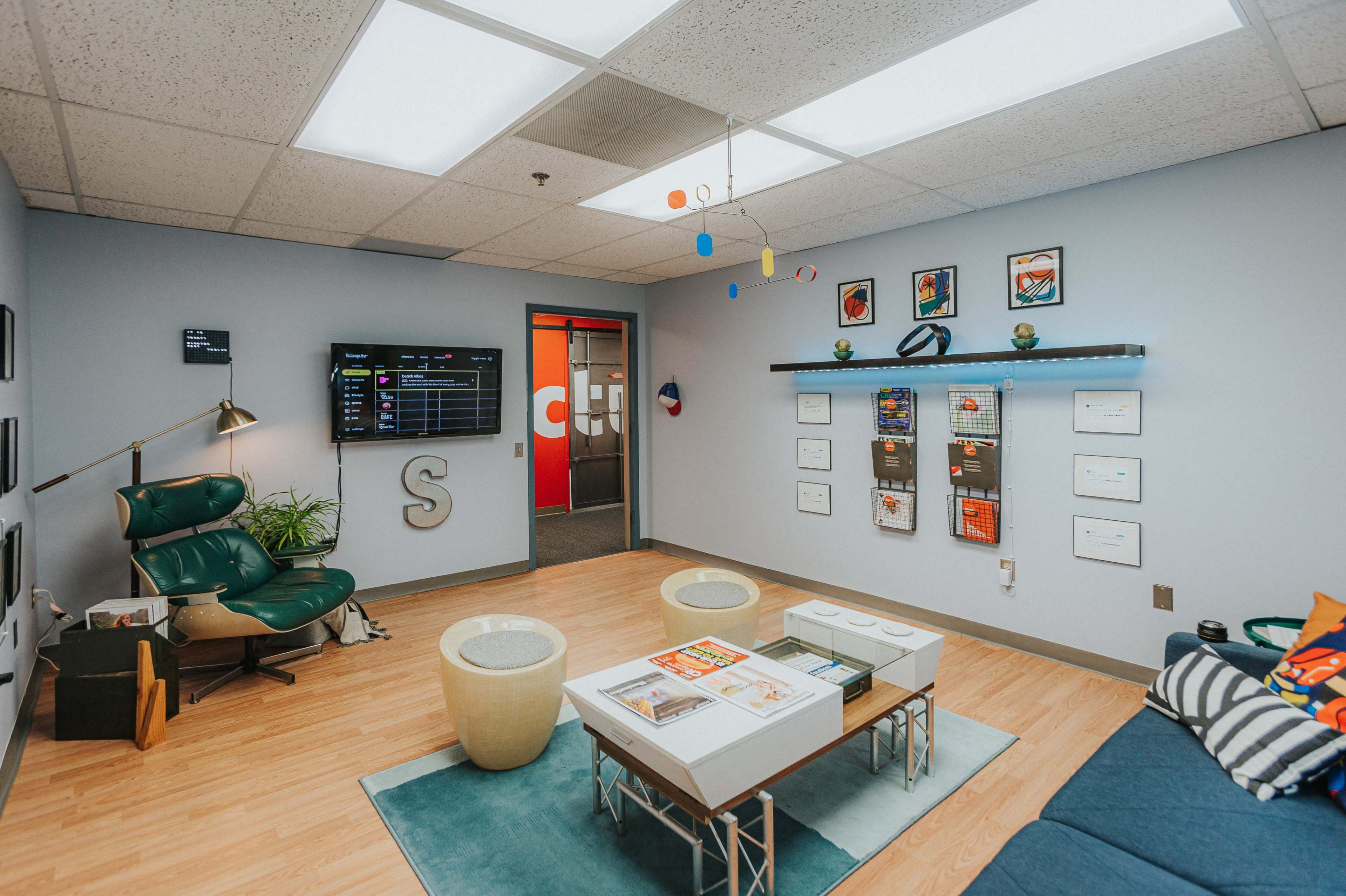 A modern office lounge area with a television on the wall, a green armchair, a low table with magazines, and wall-mounted shelves displaying framed certificates and artwork.
