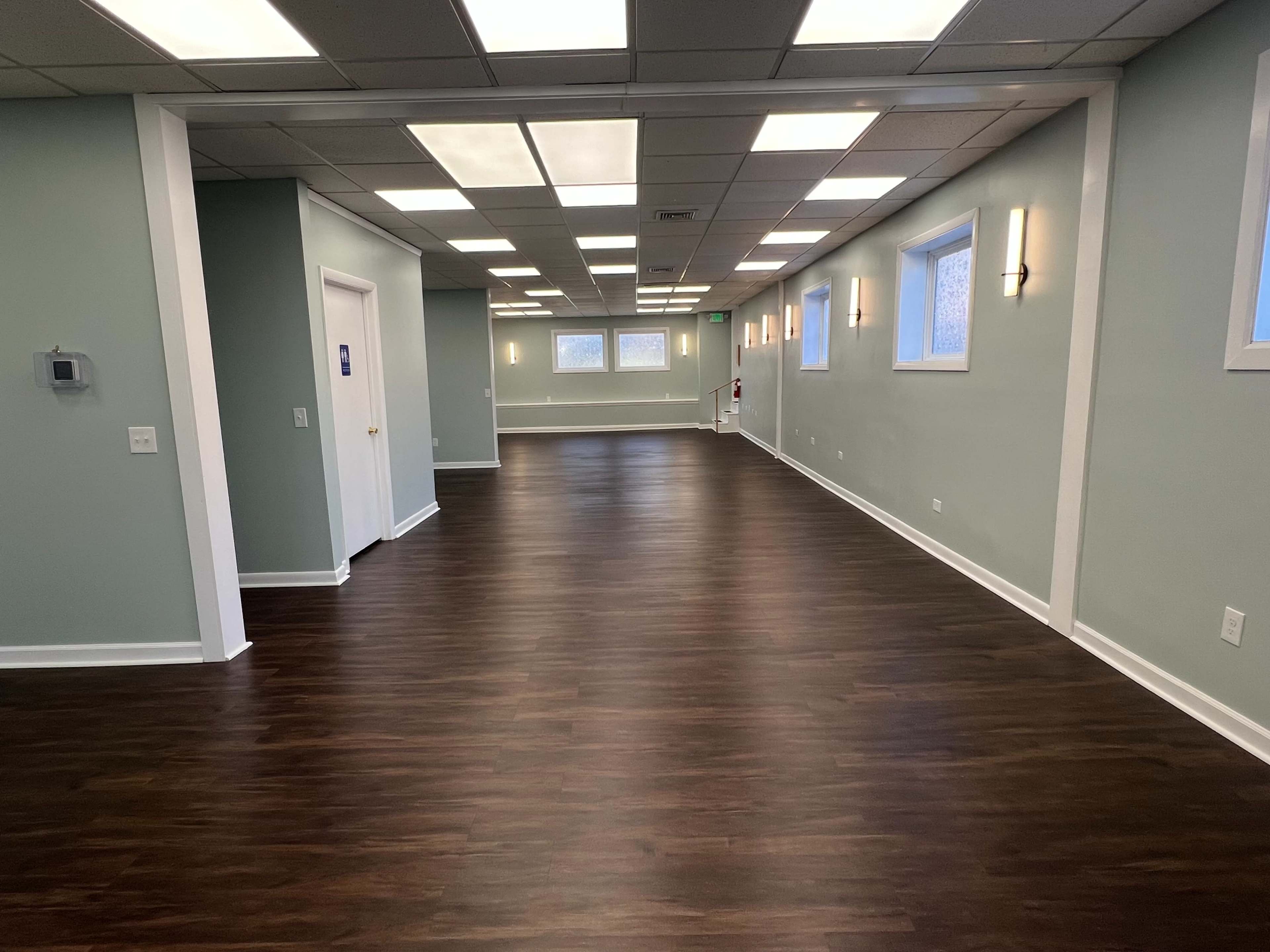 The image shows a well-lit hallway in a building with light green walls, wooden flooring, and multiple windows along one side.
