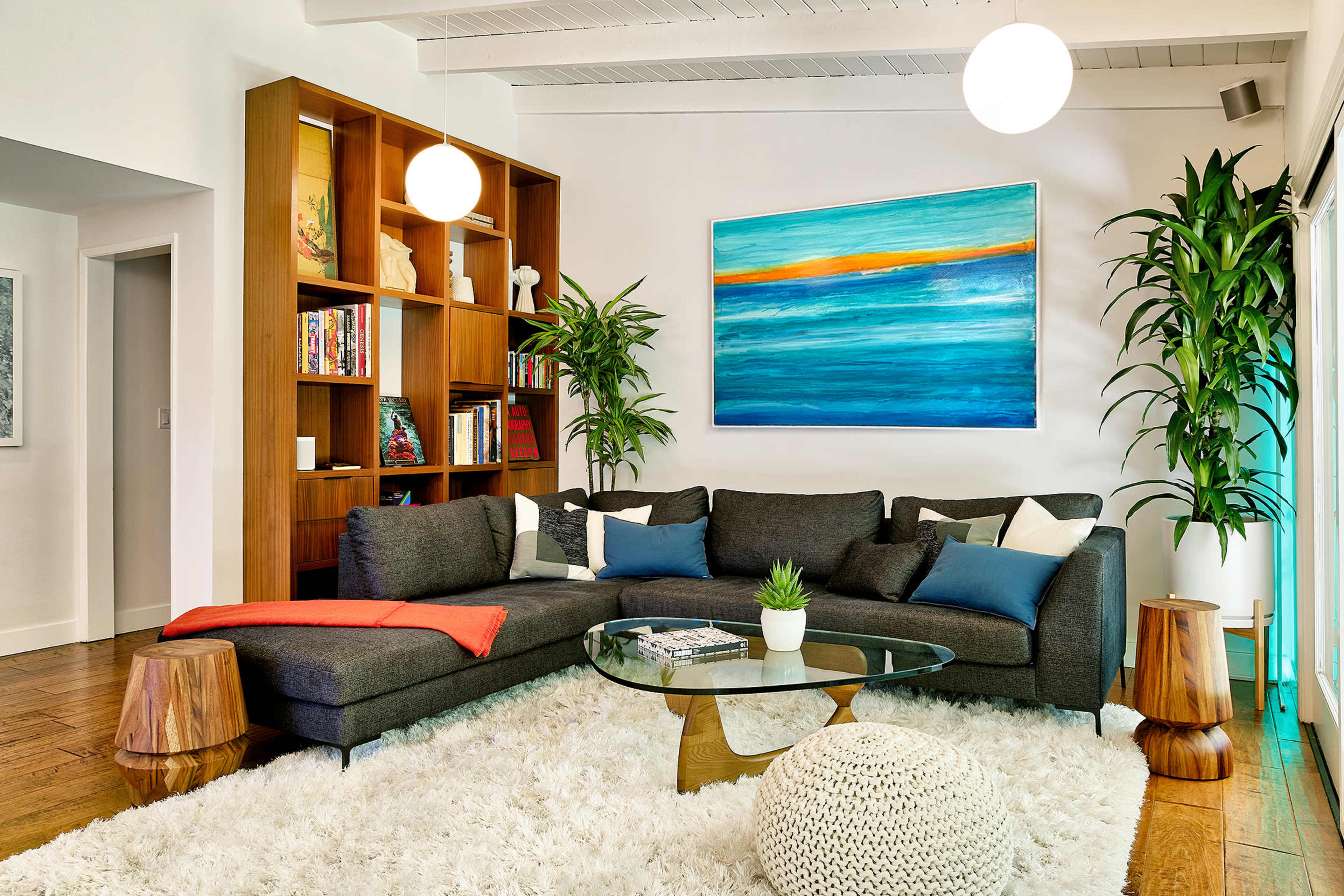 A modern living room features a sectional sofa, a circular glass coffee table, a bookshelf, and plants, with a colorful artwork hanging on the wall.