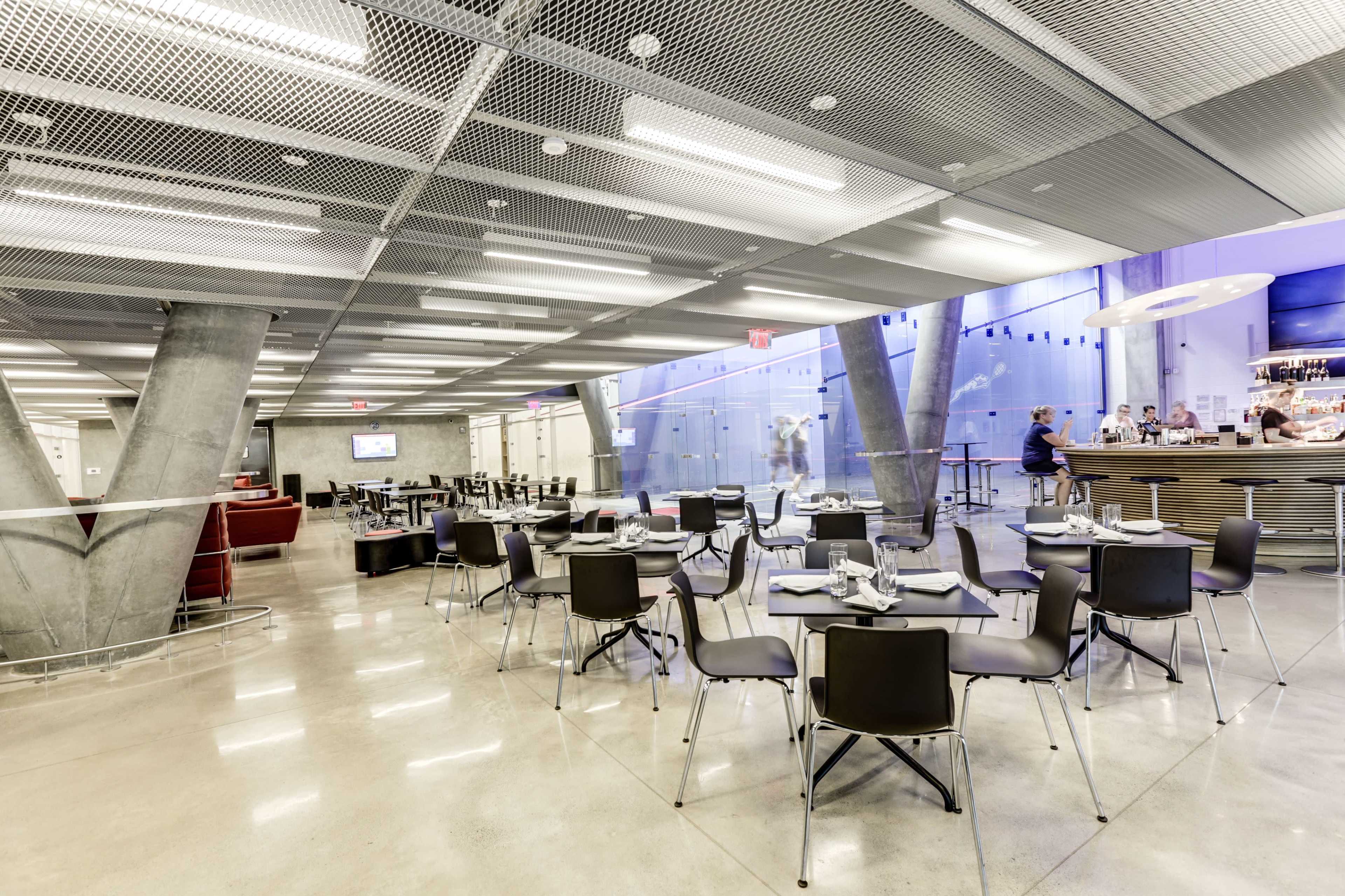 The image shows a modern dining area with sleek furniture, exposed concrete structures, and a bar along one side, illuminated by blue lighting.