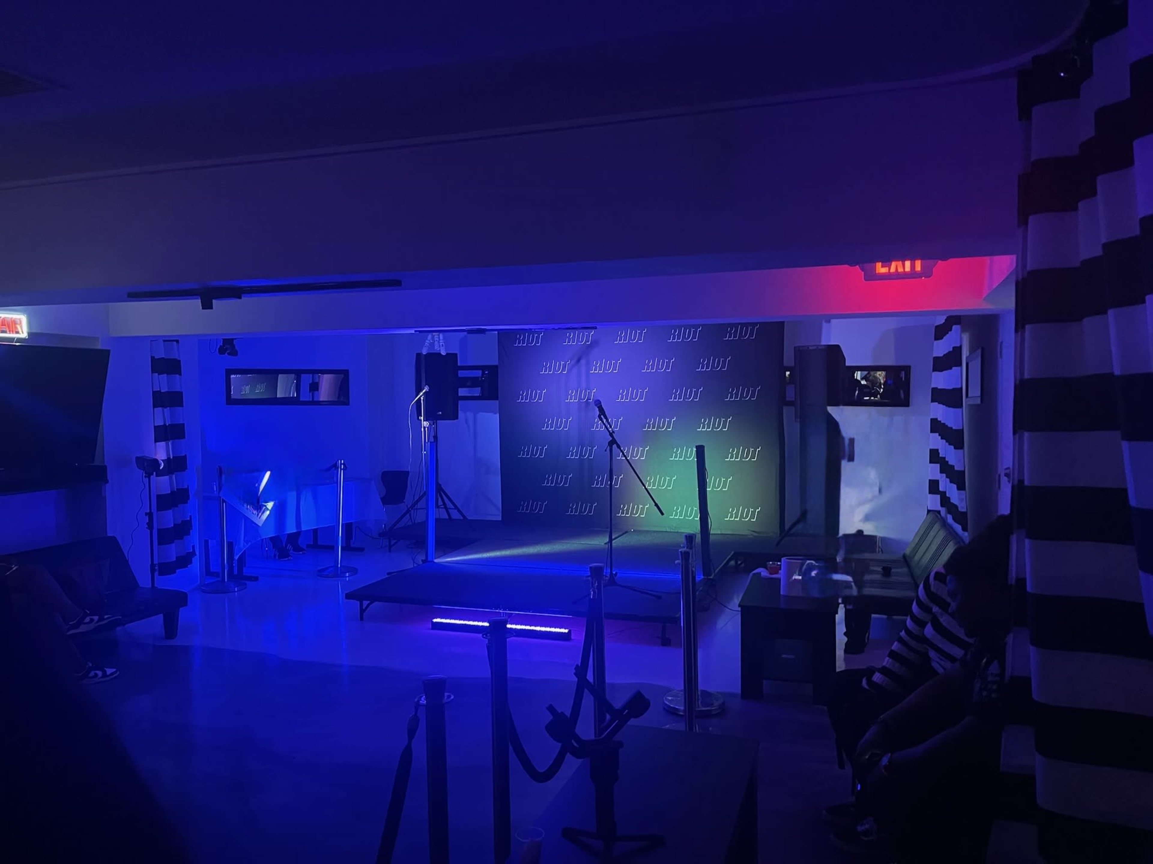 A dimly lit event space features a stage with a backdrop and colorful lighting, surrounded by chairs and a lounge area.