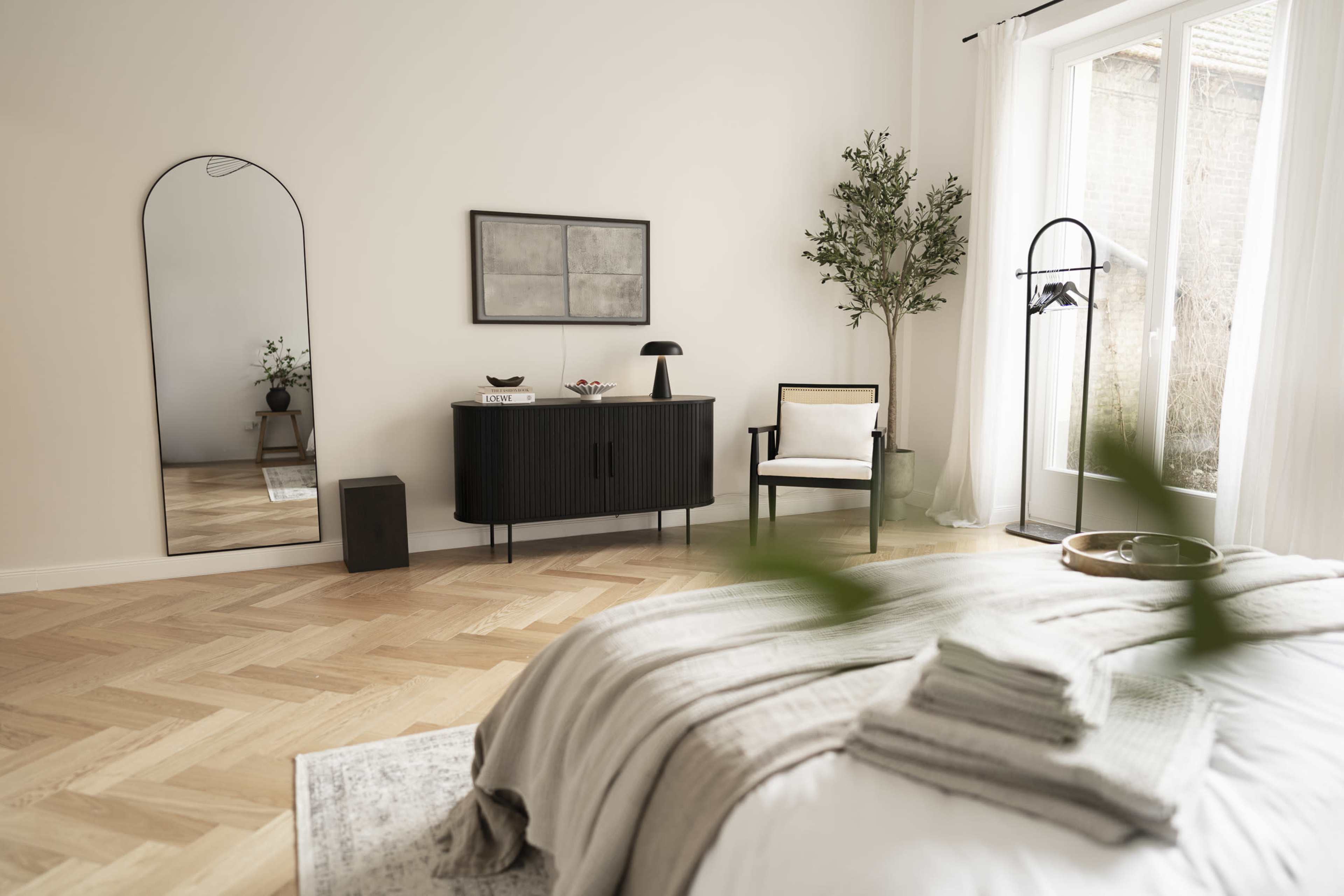 A minimalist bedroom features a large mirror, a dark wood cabinet, and a chair beside a window with natural light filtering in.