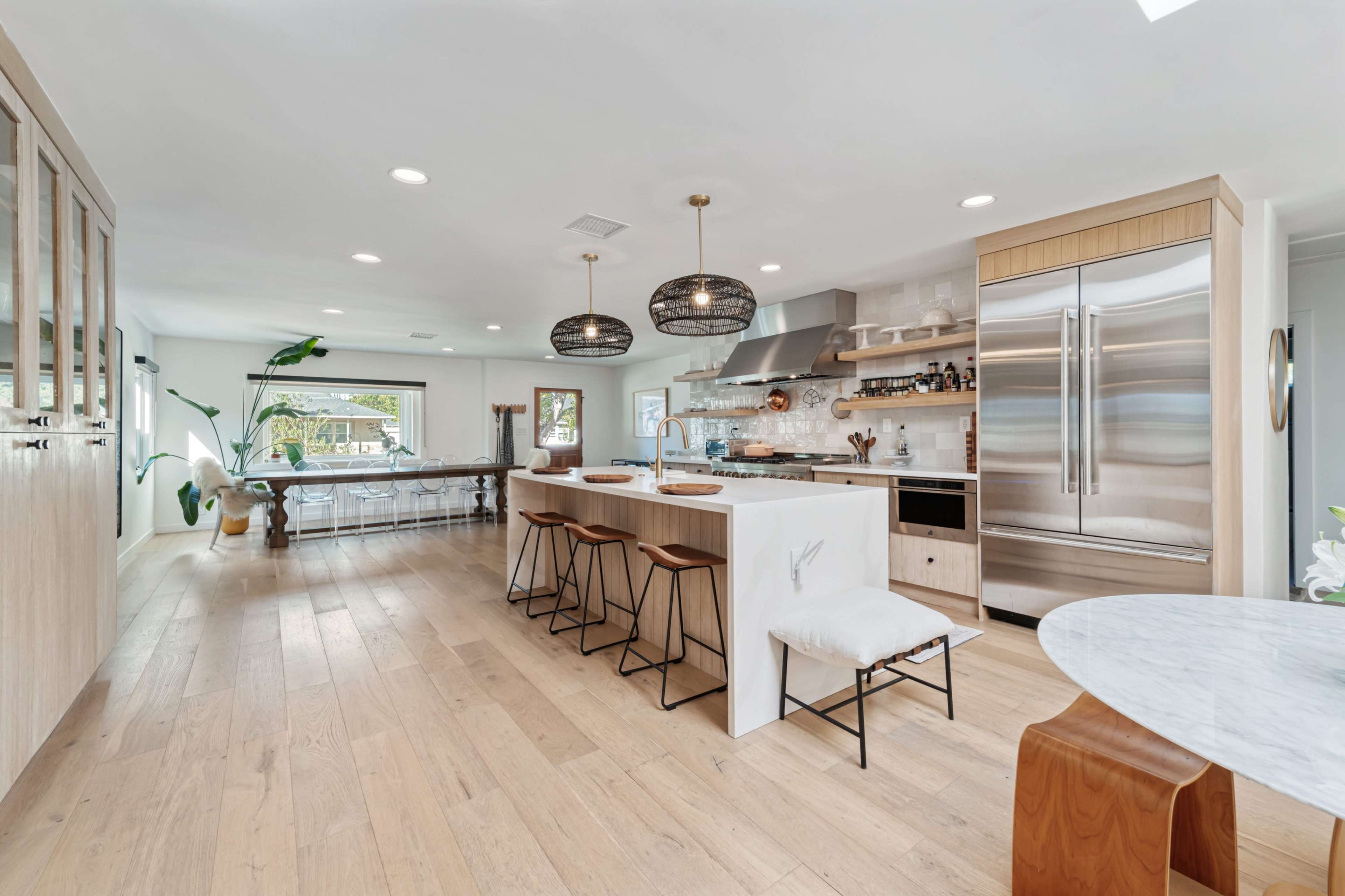 The image shows a modern kitchen with a large central island, bar stools, stainless steel appliances, and a dining area in the background.