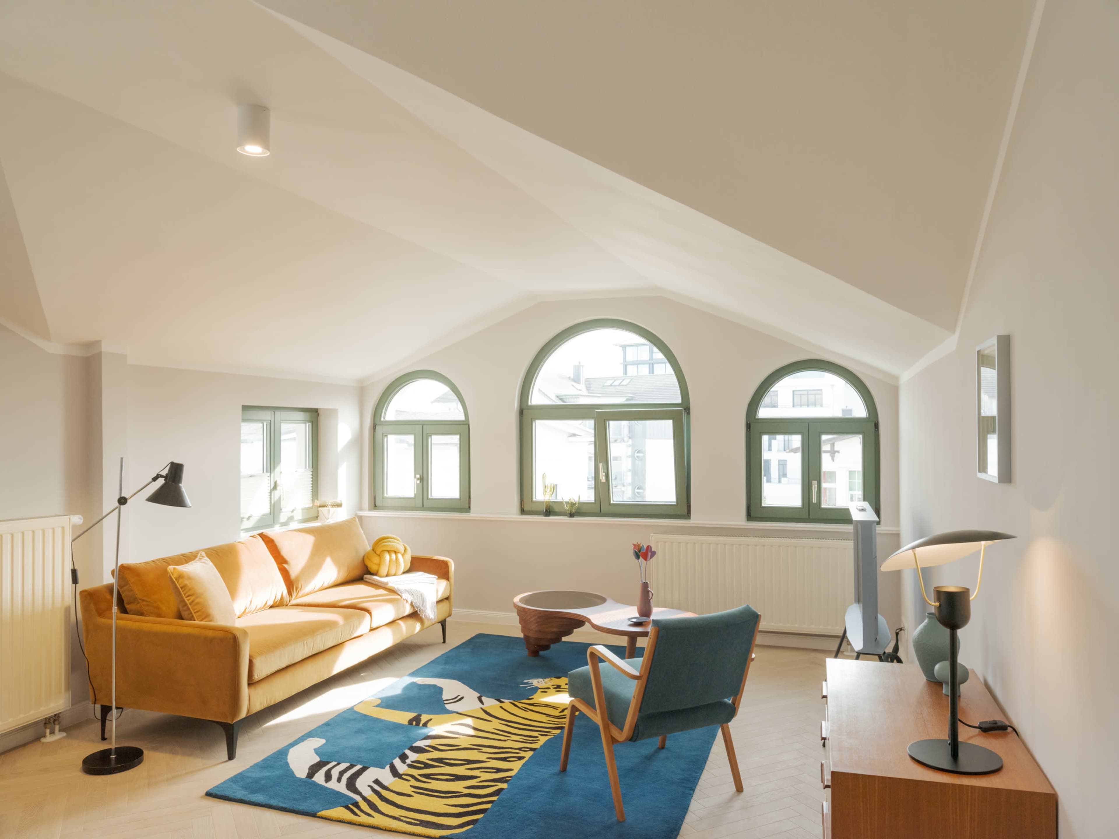 The image shows a modern living room with a yellow sofa, teal chairs, and a large blue rug featuring a tiger design, illuminated by natural light through arched windows.
