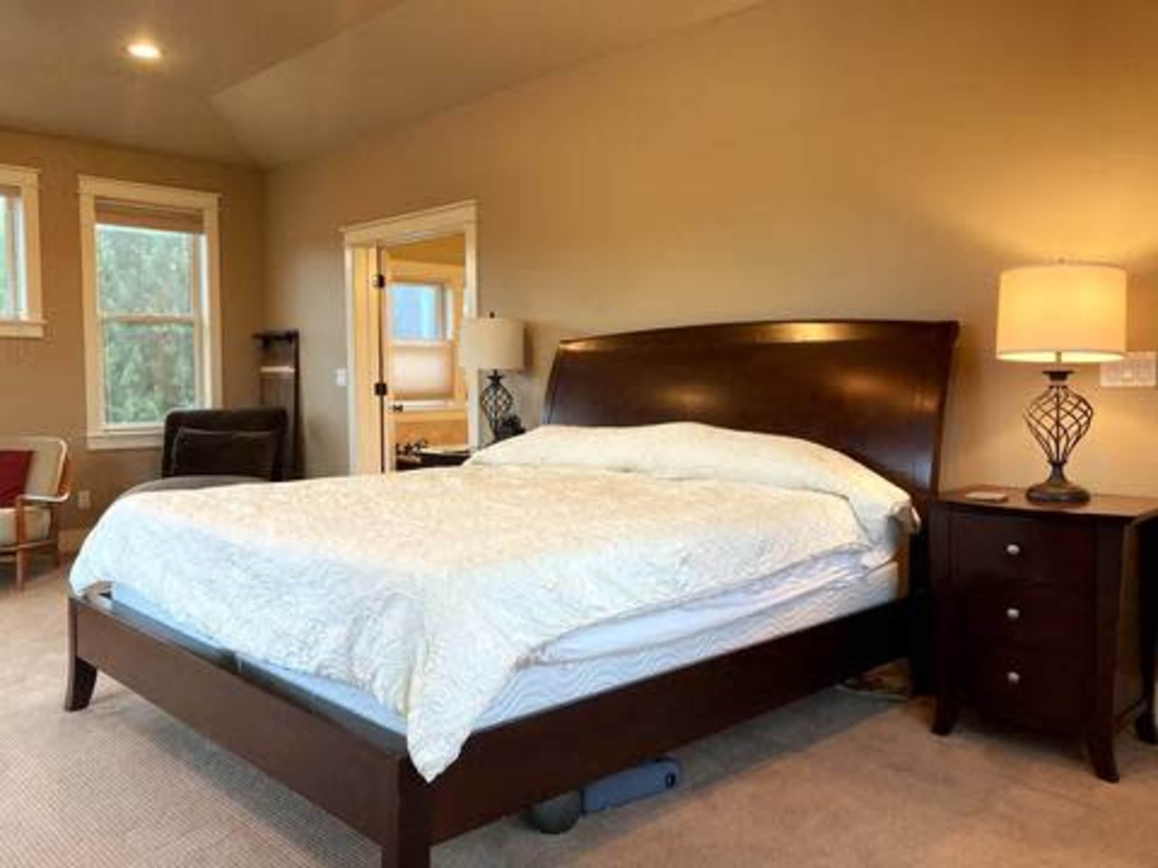 A spacious bedroom featuring a large wooden bed with a light-colored bedspread, a nightstand with a lamp, and windows allowing natural light.