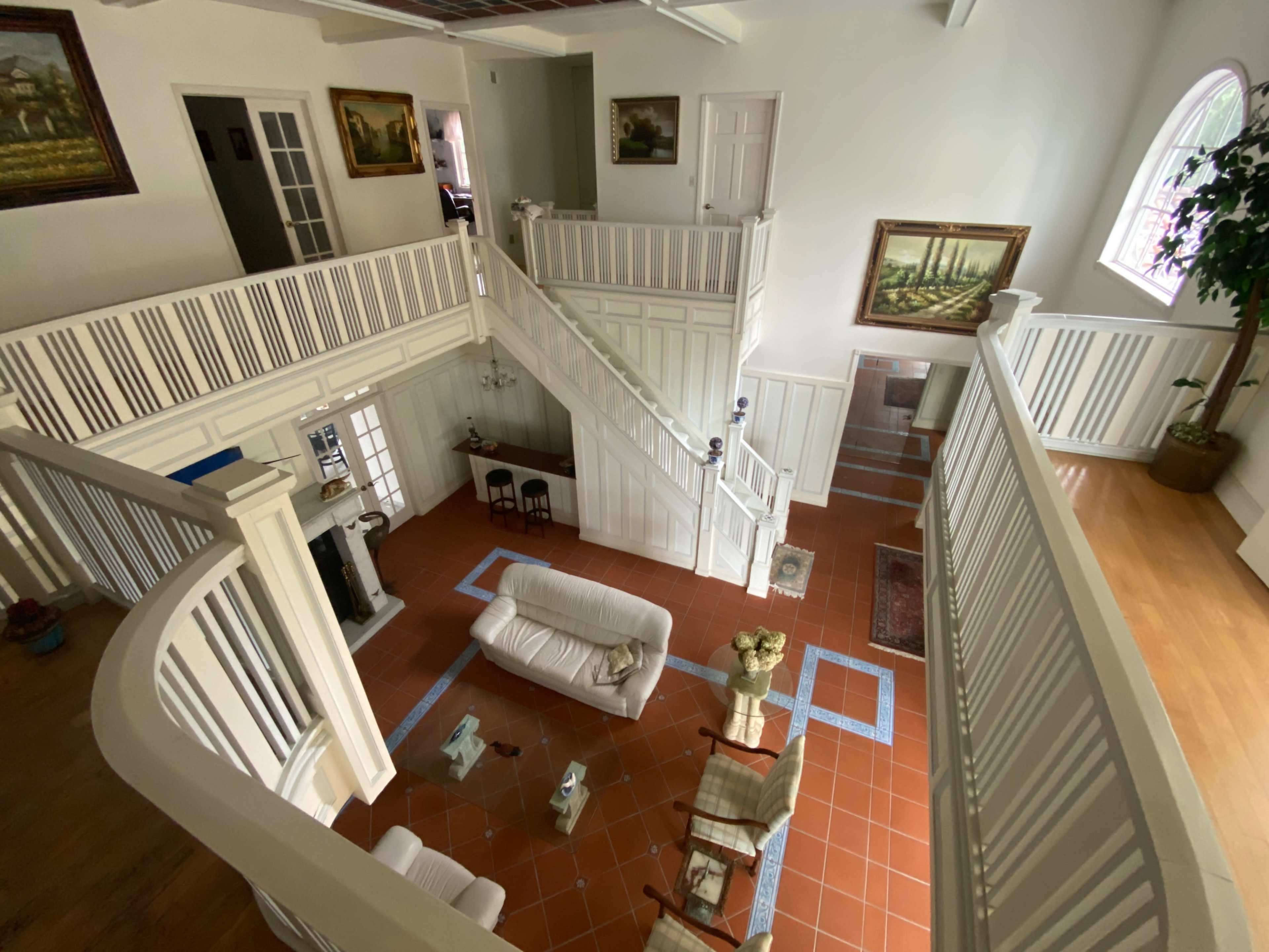 The image shows a spacious, multi-level interior of a house with white wooden railings, a tiled floor, and various pieces of furniture, including a couch and chairs.