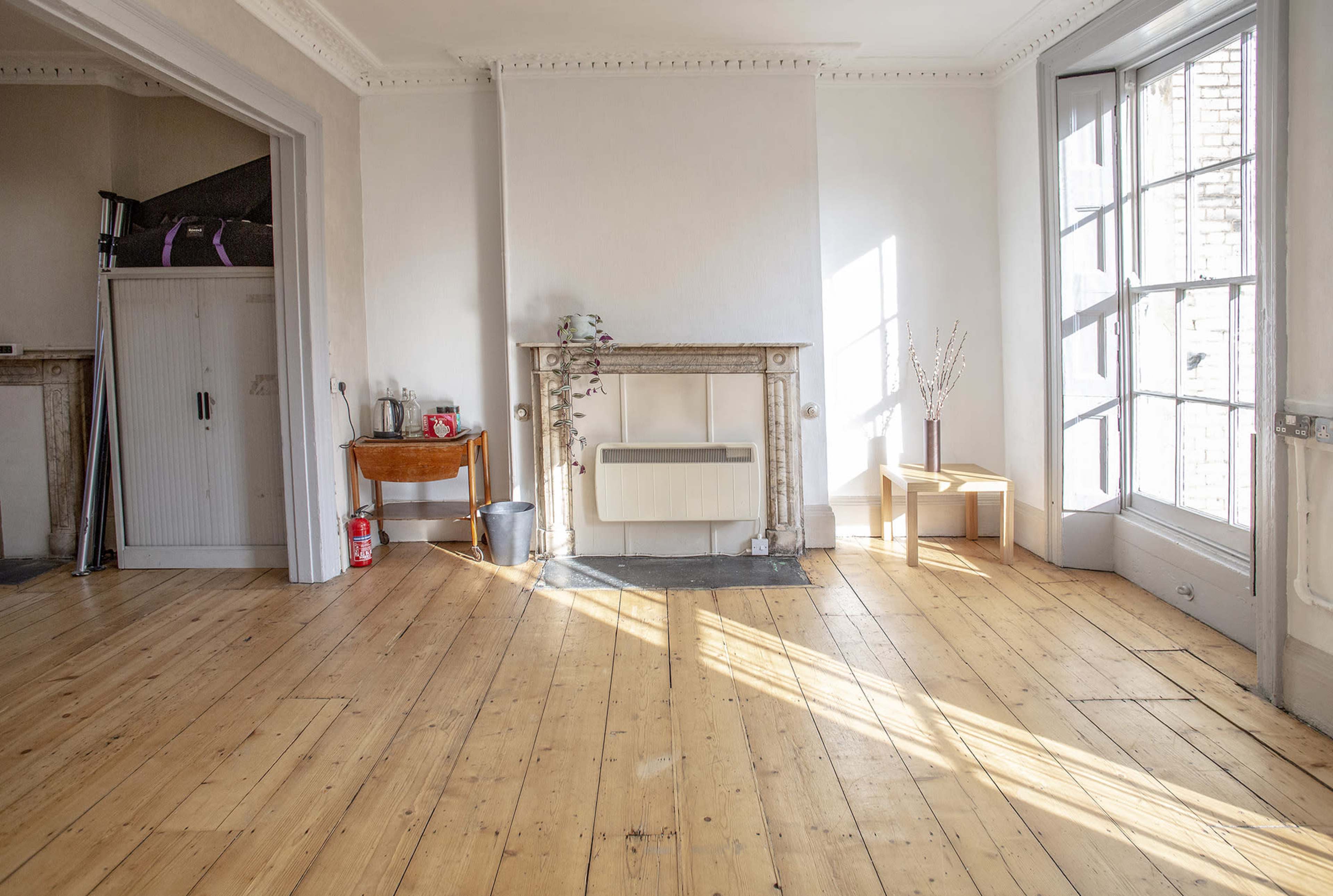 A bright room featuring wooden floors, large windows, and minimal furnishings, including a small table and a decorative mantelpiece.