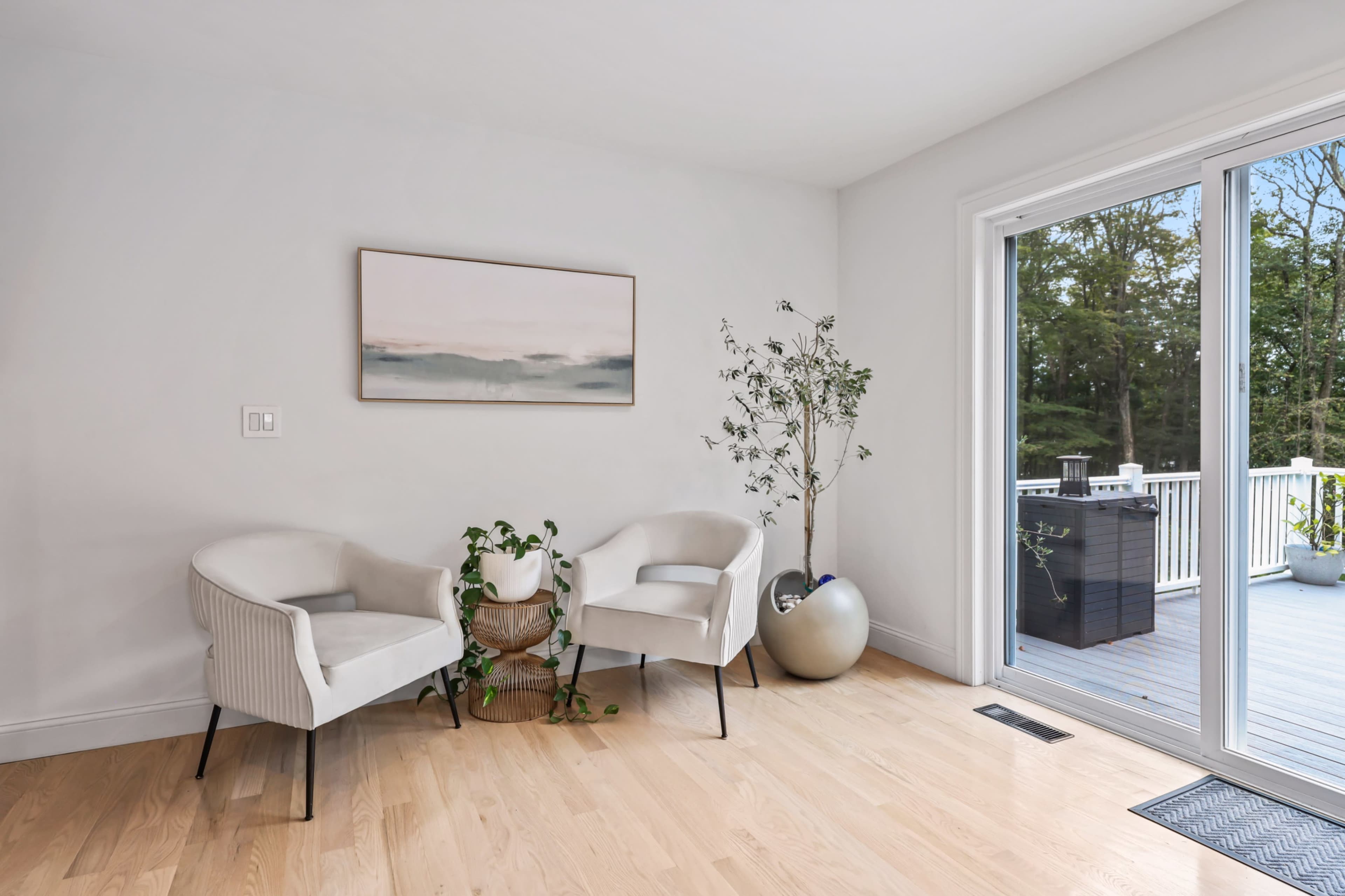 The image shows a minimalist interior space with two white armchairs, a small plant in a decorative pot, and a large sliding glass door opening to a wooden deck.