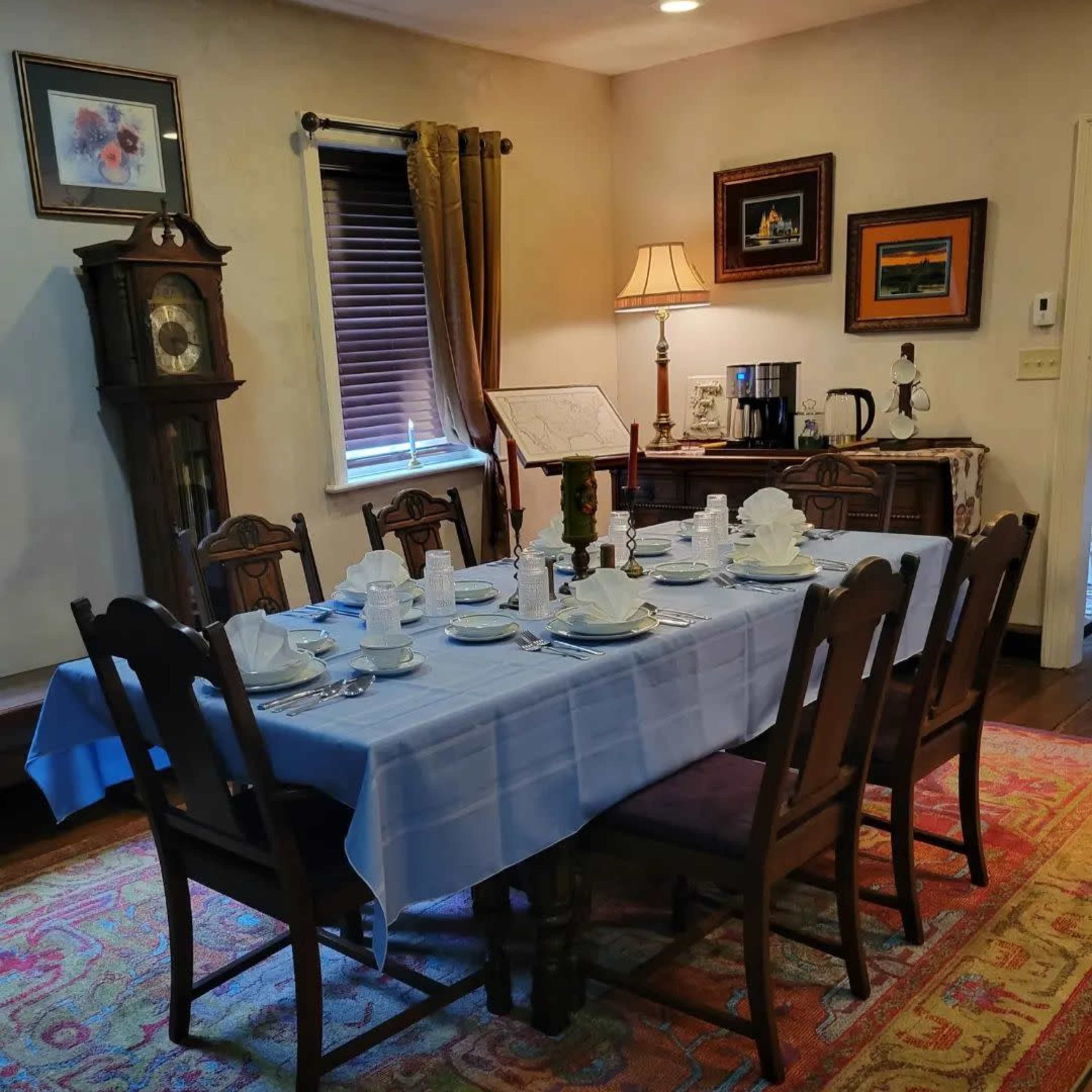 A dining room is set with a blue tablecloth and a table arranged with white dishware, surrounded by wooden chairs and decorated with various artworks.