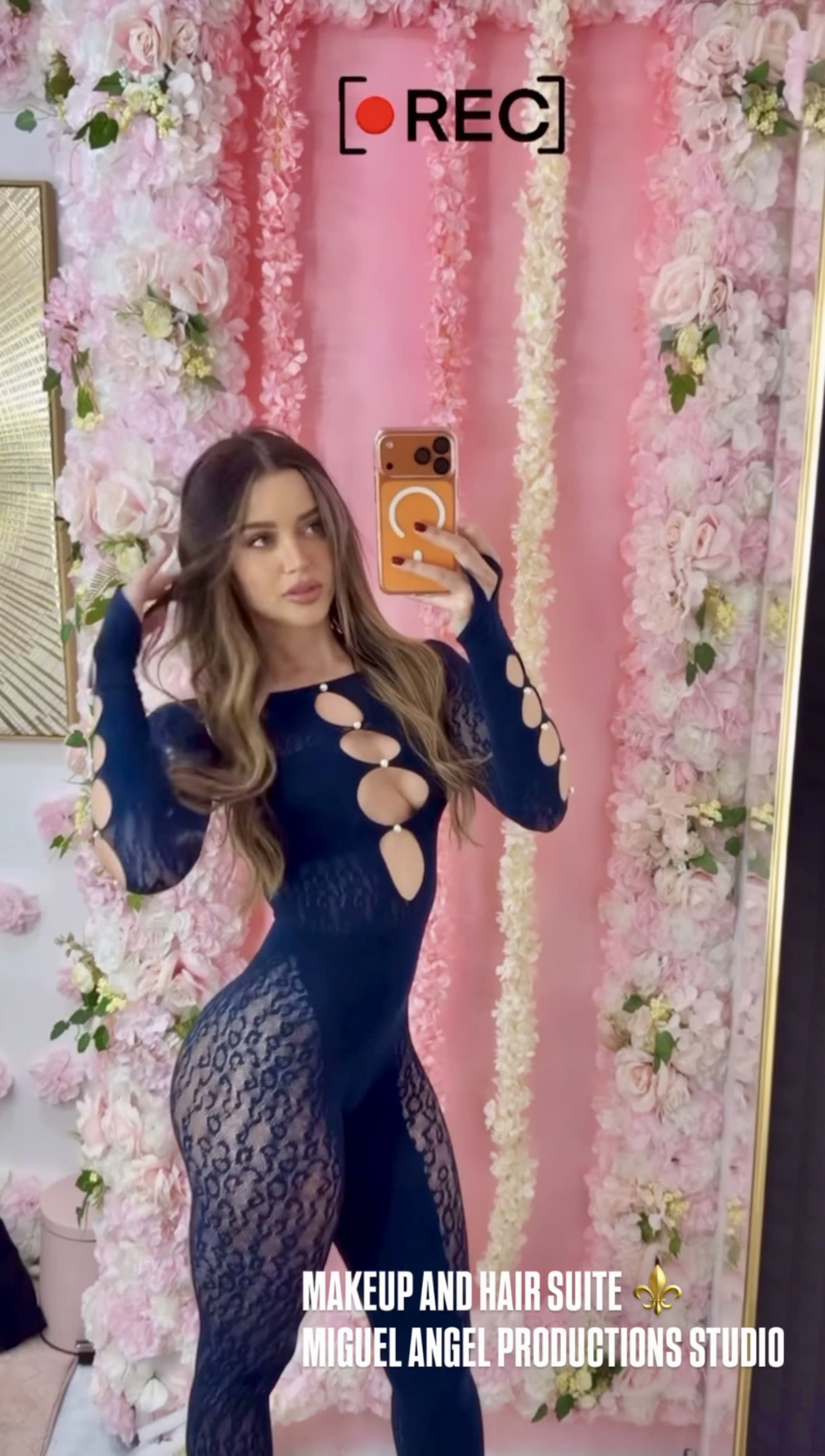 A woman poses in front of a floral backdrop while taking a selfie in a fitted, patterned outfit.