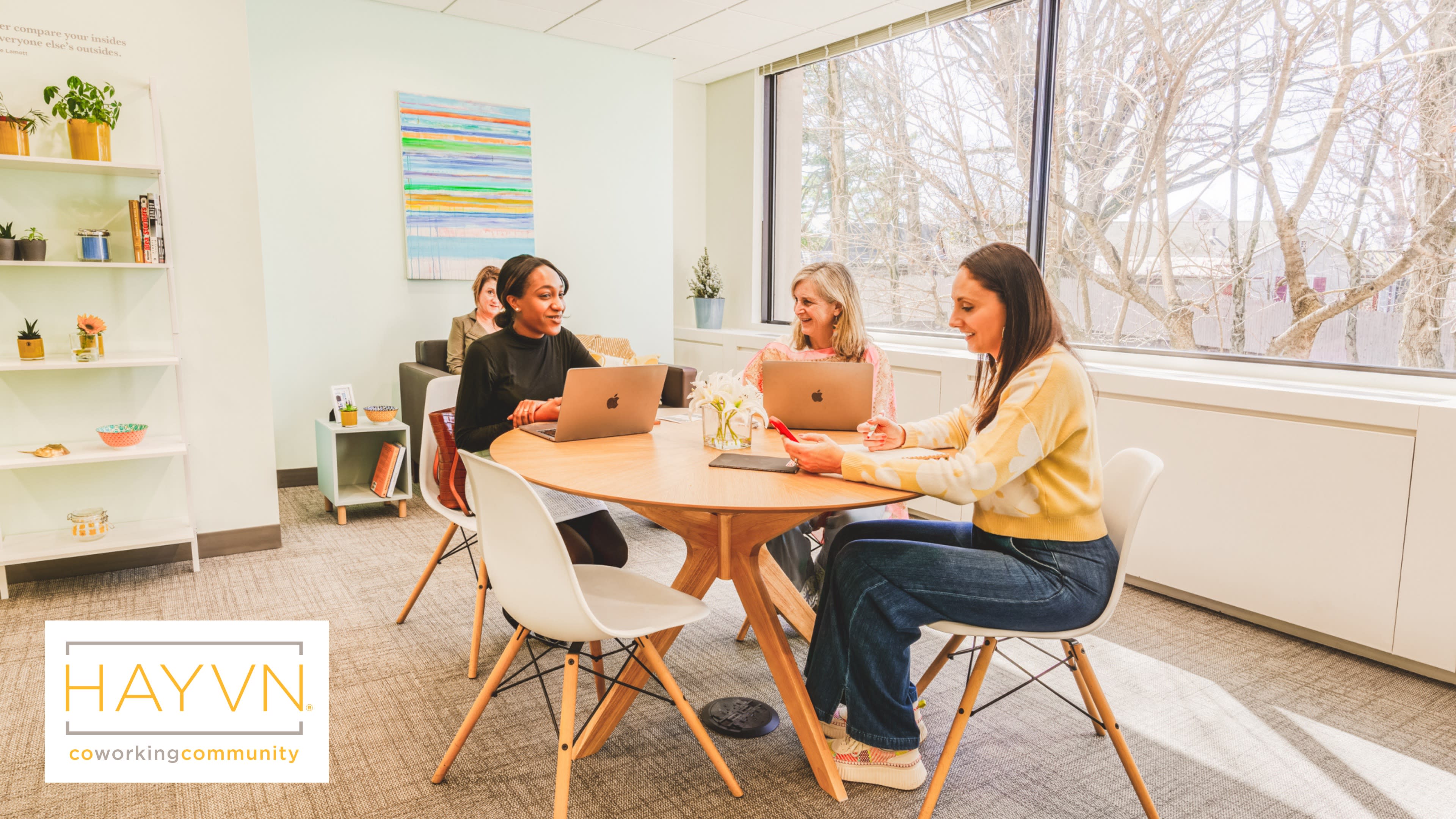 Four women are seated around a wooden table in a well-lit coworking space, each using laptops and engaged in a discussion.