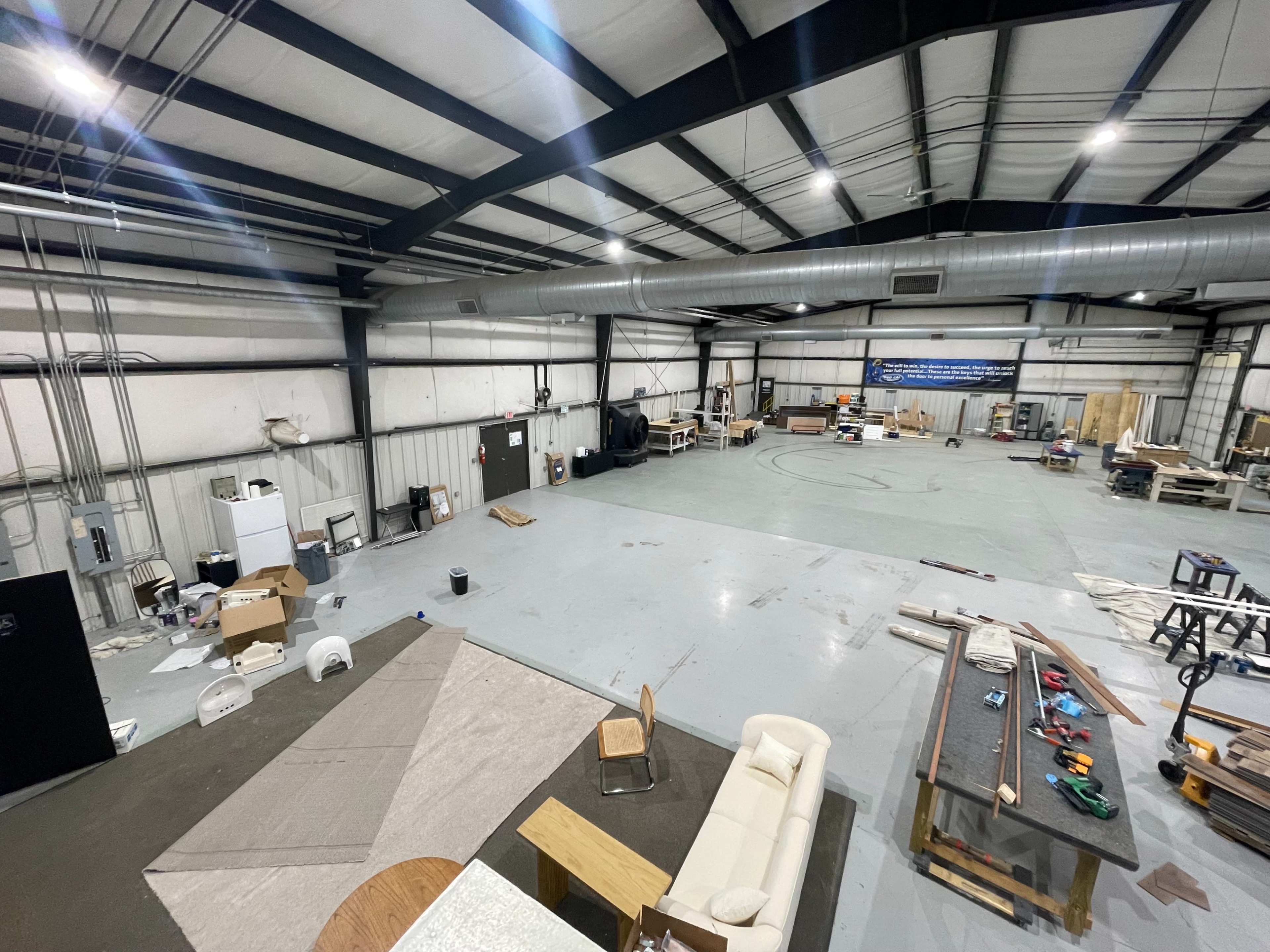 The image shows a spacious industrial workspace with various tools and materials strewn around, including furniture, equipment, and unfinished projects.