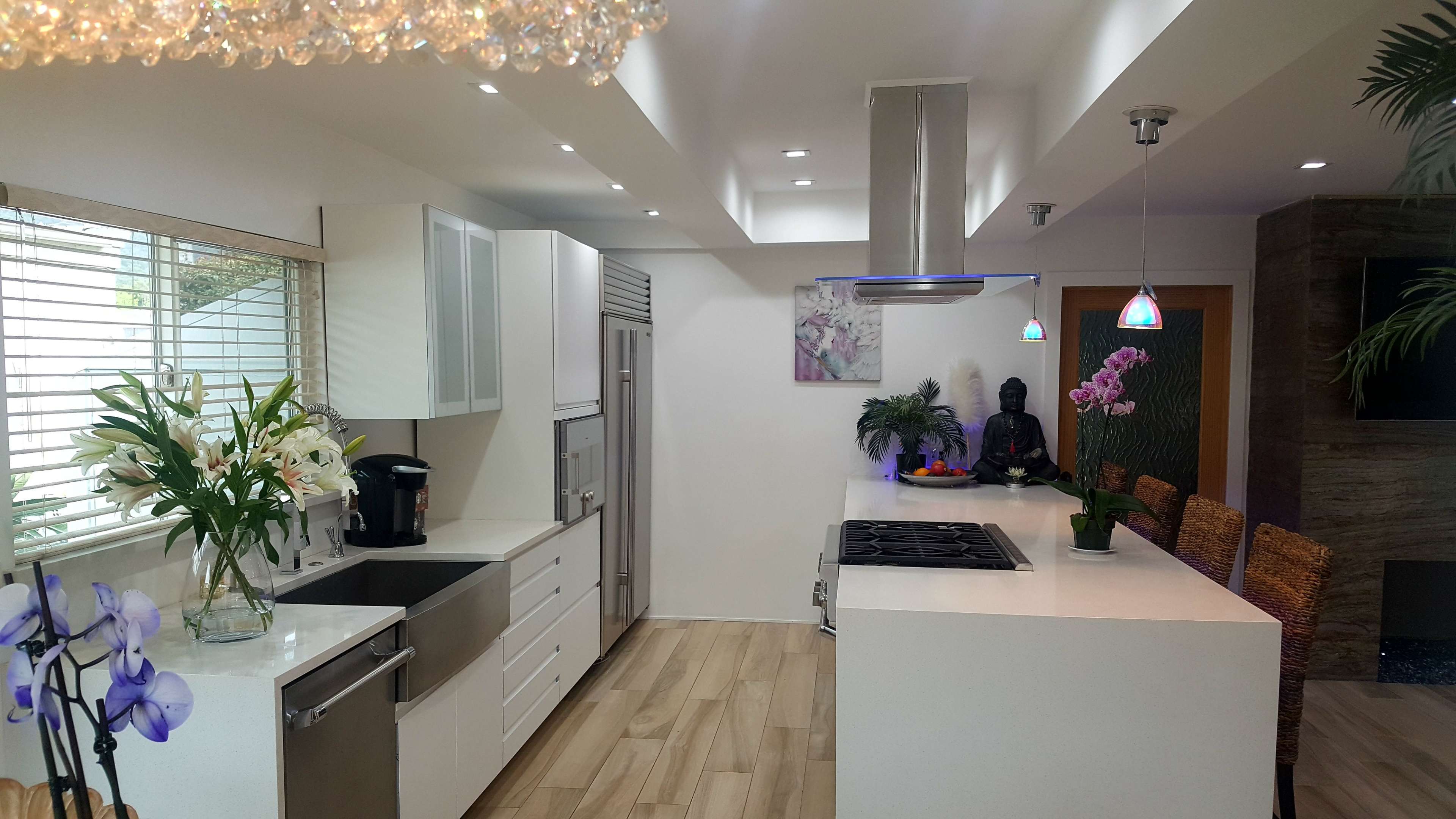The image shows a modern kitchen with white cabinetry, a central island with a stovetop, and a dining area featuring wicker chairs and decorative plants.