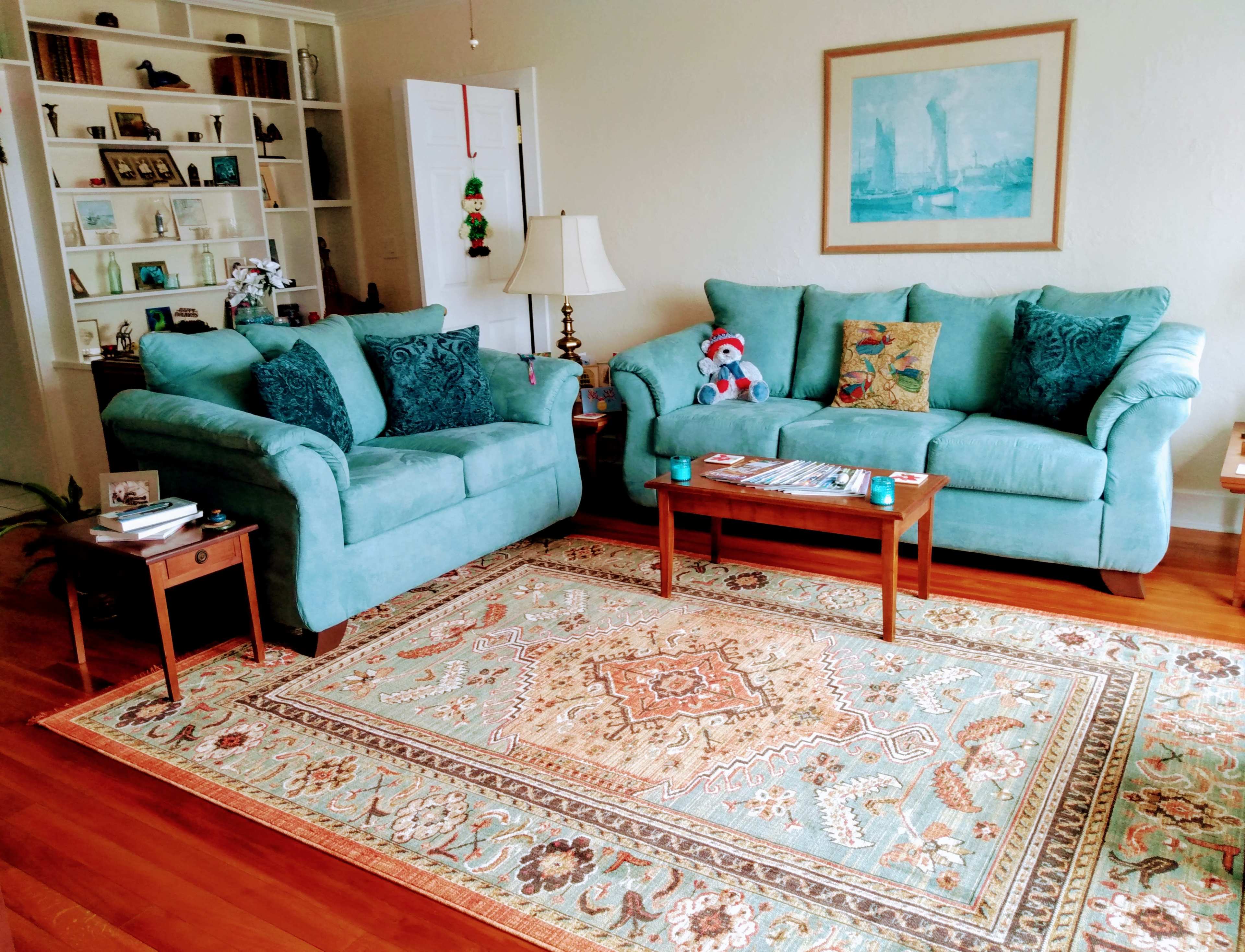 A light-colored living room features two turquoise sofas, a wooden coffee table, and a decorative rug, with built-in shelves in the background.
