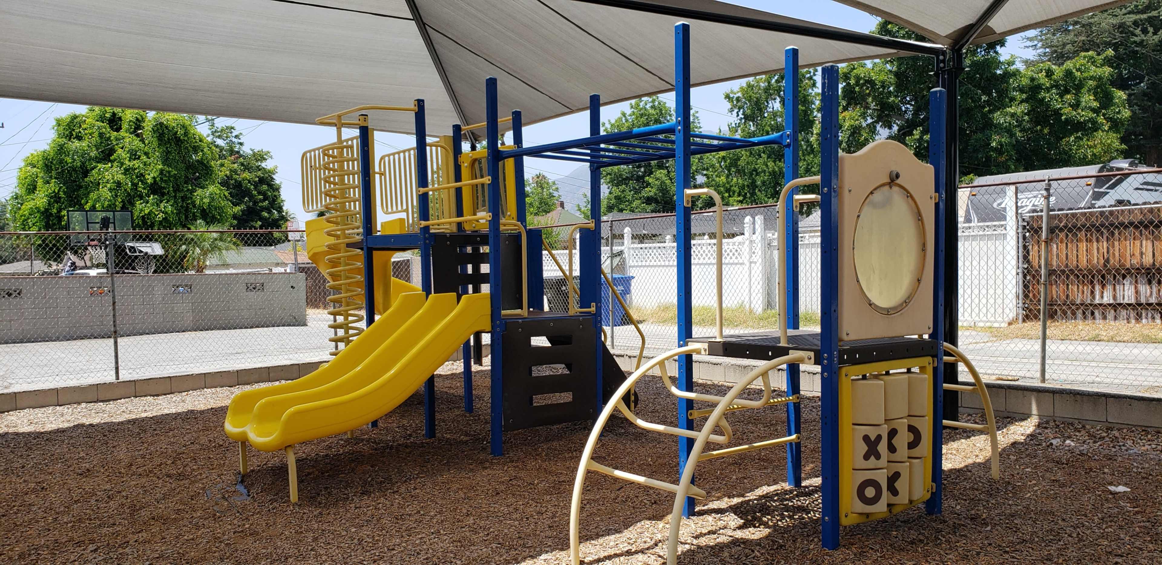 The image shows a playground with a yellow slide, climbing structures, and a large spiral slide under a shaded canopy.