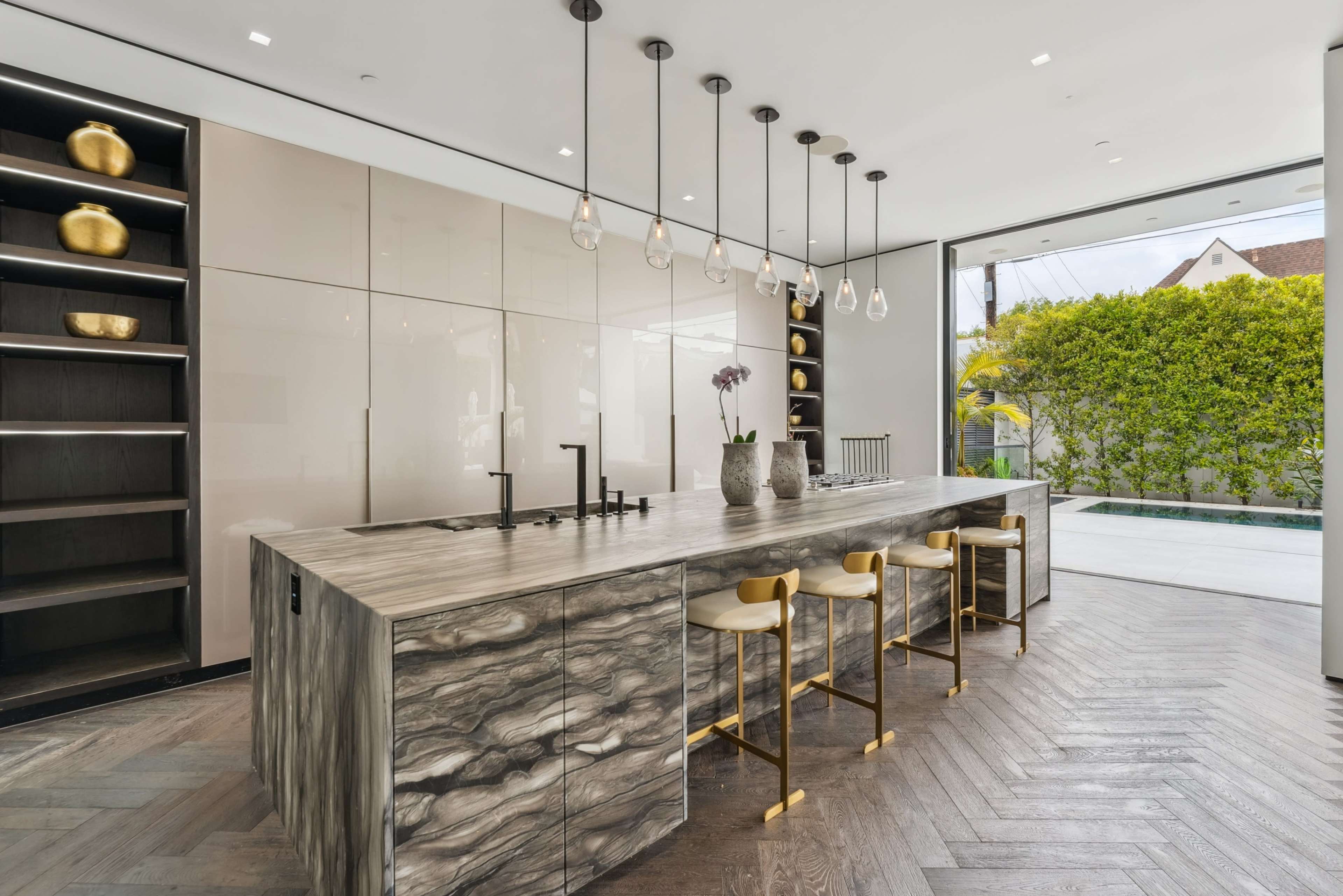 The image shows a modern kitchen with a large island featuring a marbled countertop, bar stools, pendant lighting, and large windows that offer a view of greenery outside.