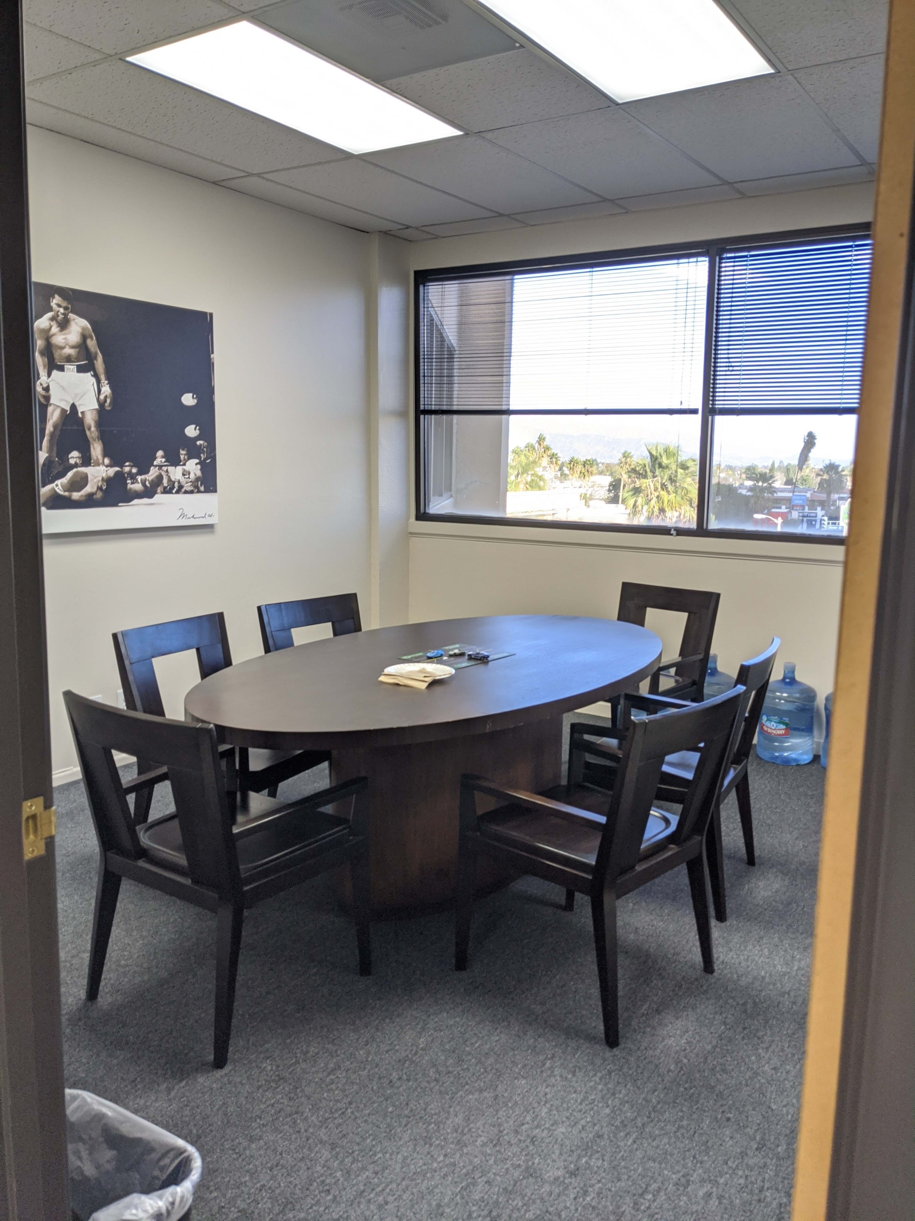 A conference room features an oval table surrounded by seven dark chairs, with a window displaying a view outside and a water dispenser in the corner.