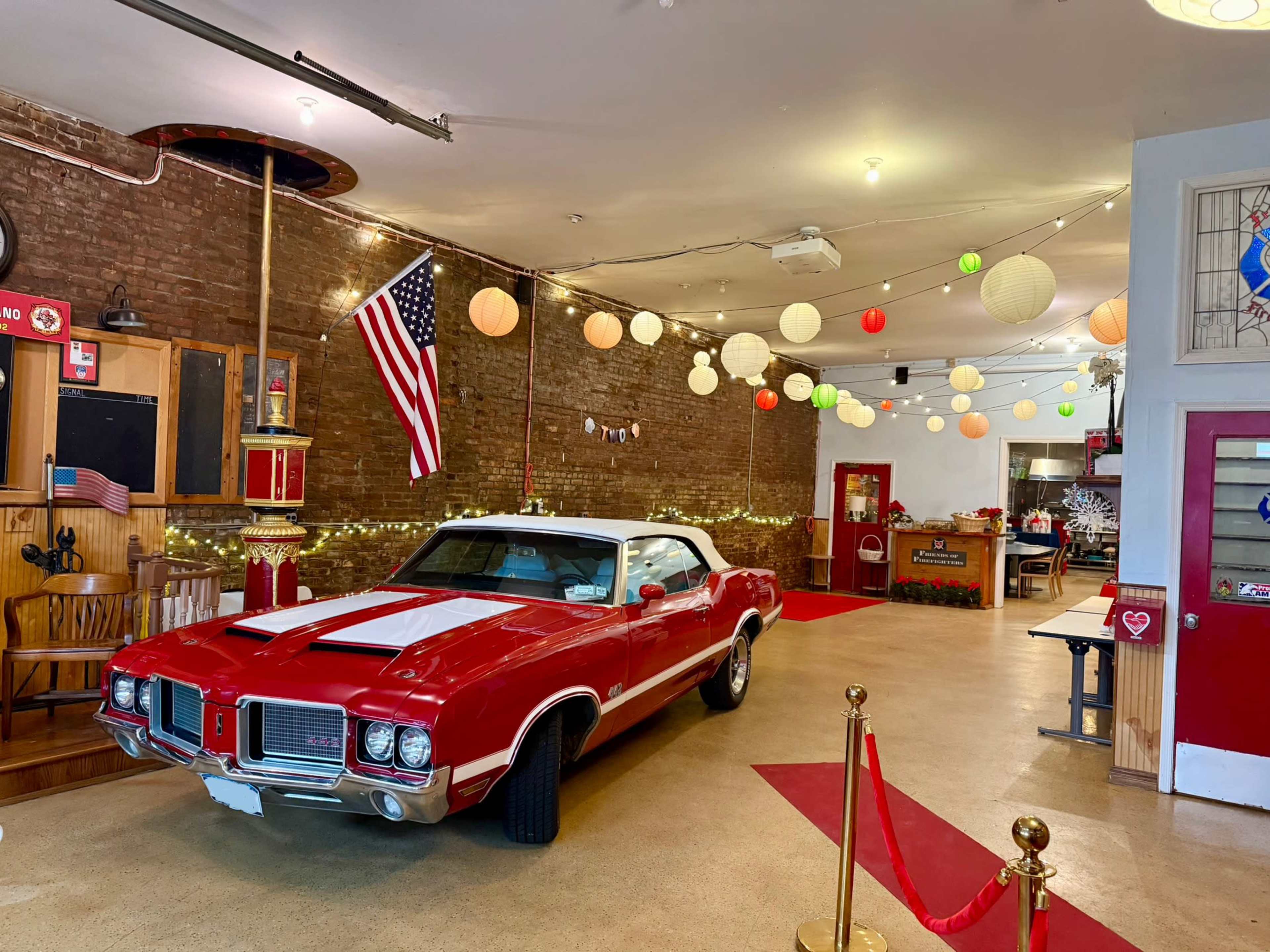 The image shows a vintage red car parked inside a room with brick walls, a string of colorful lanterns, and a hanging American flag.
