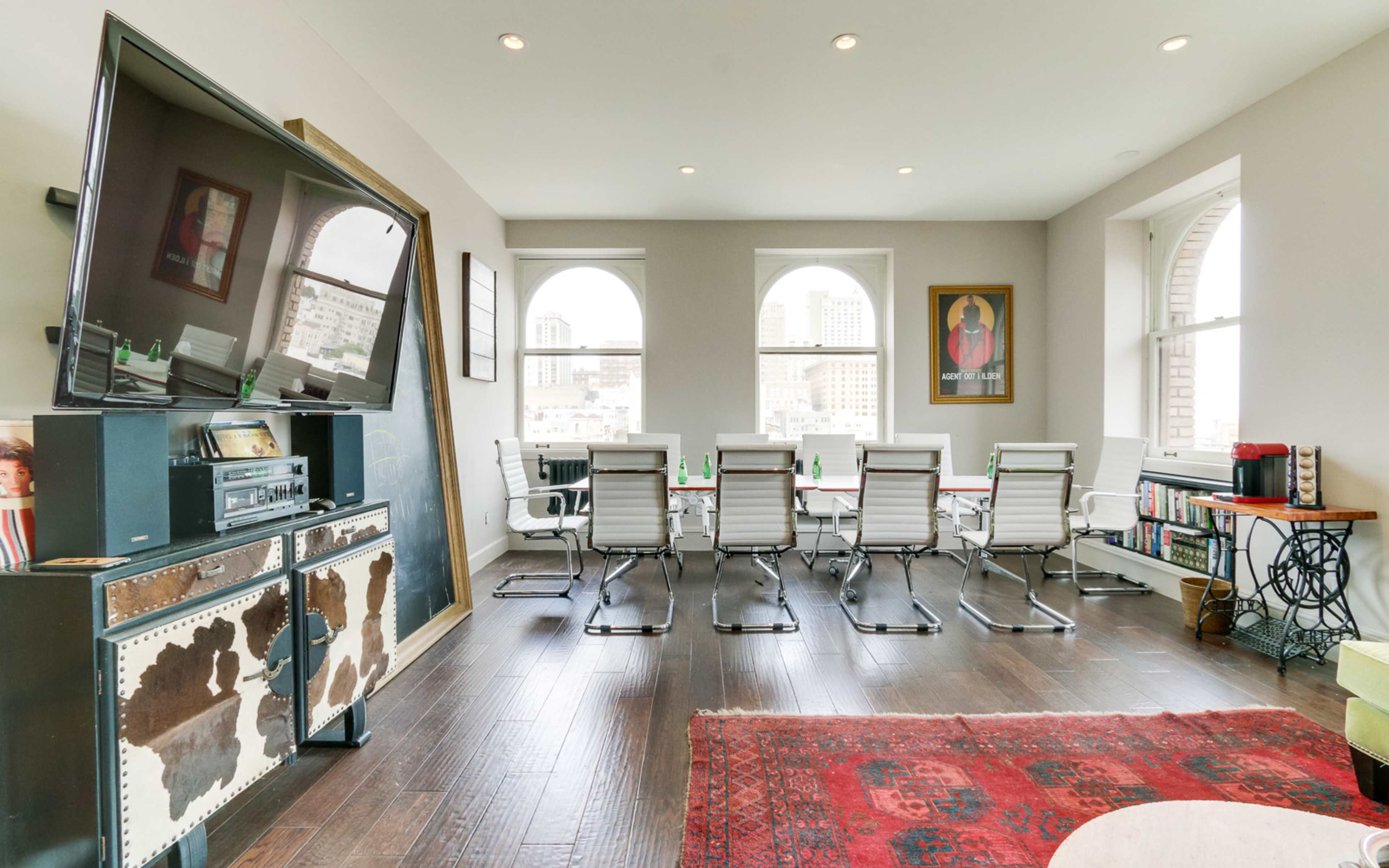 A modern conference room with a long table and several chairs, featuring large windows that provide a view of a city skyline.