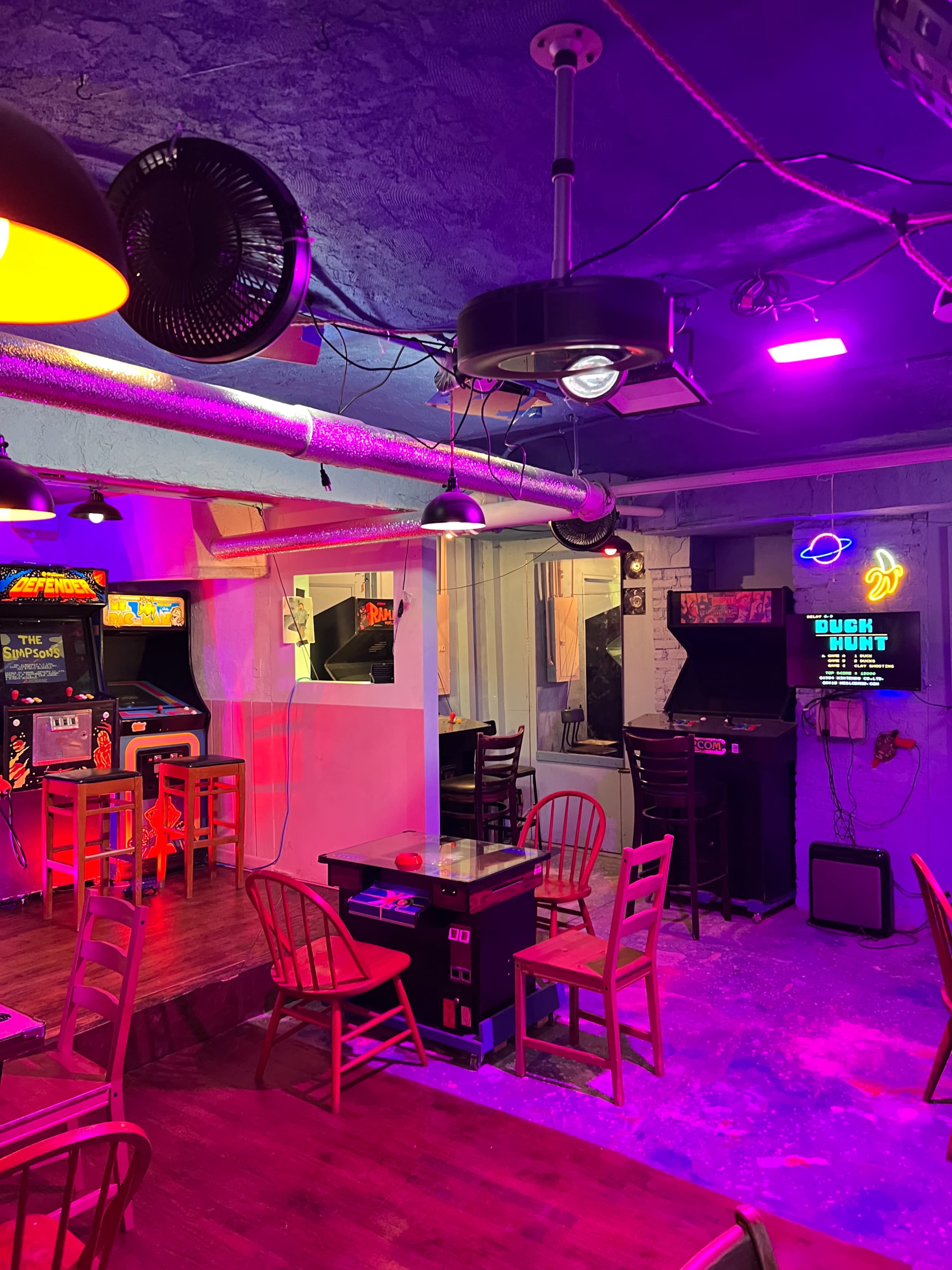 A retro arcade environment with arcade machines, wooden chairs, and colorful lighting on the walls and ceiling.