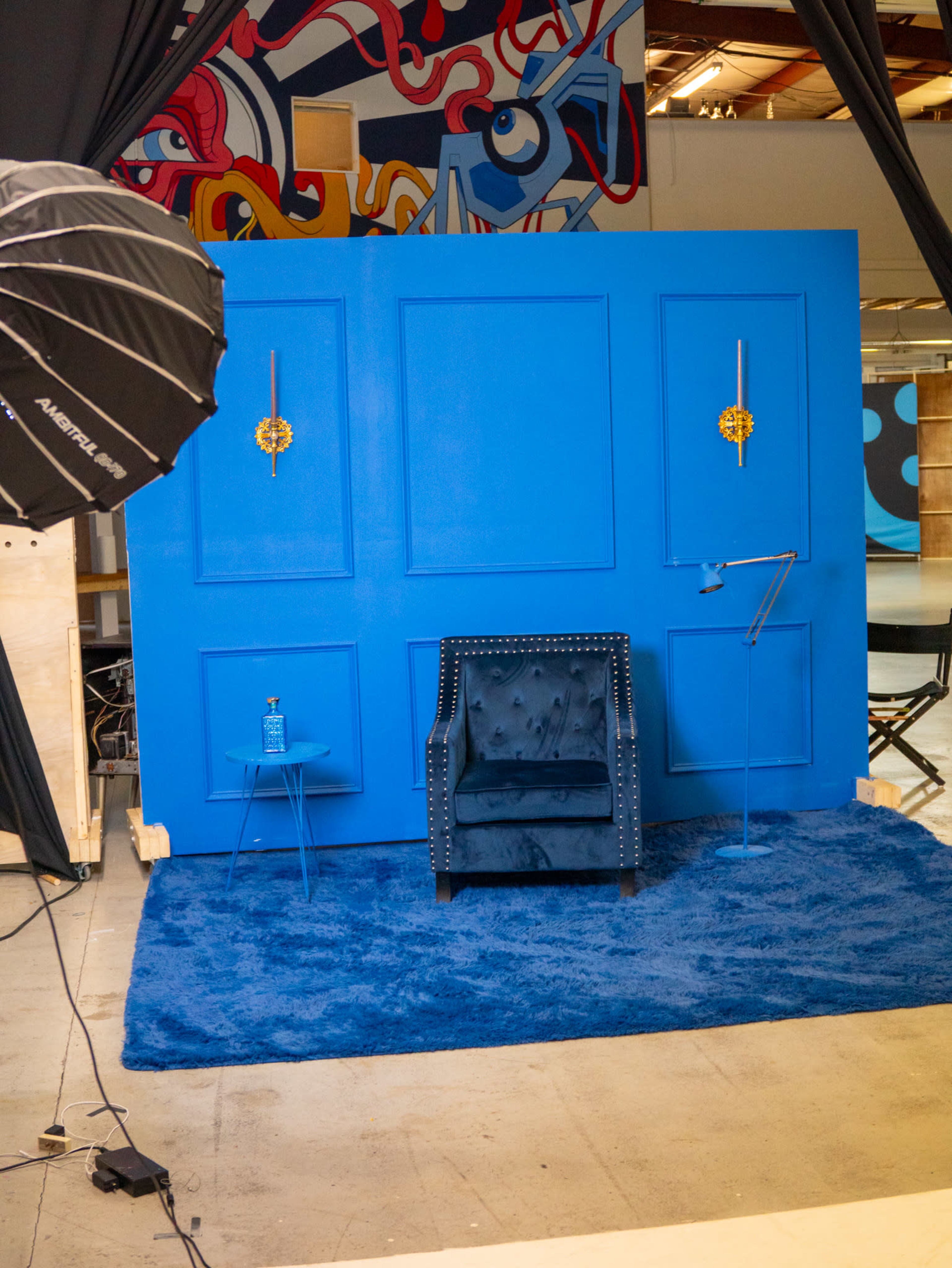 The image features a bright blue backdrop with a chair, a small table, and a floor mat, all arranged in a studio setting.