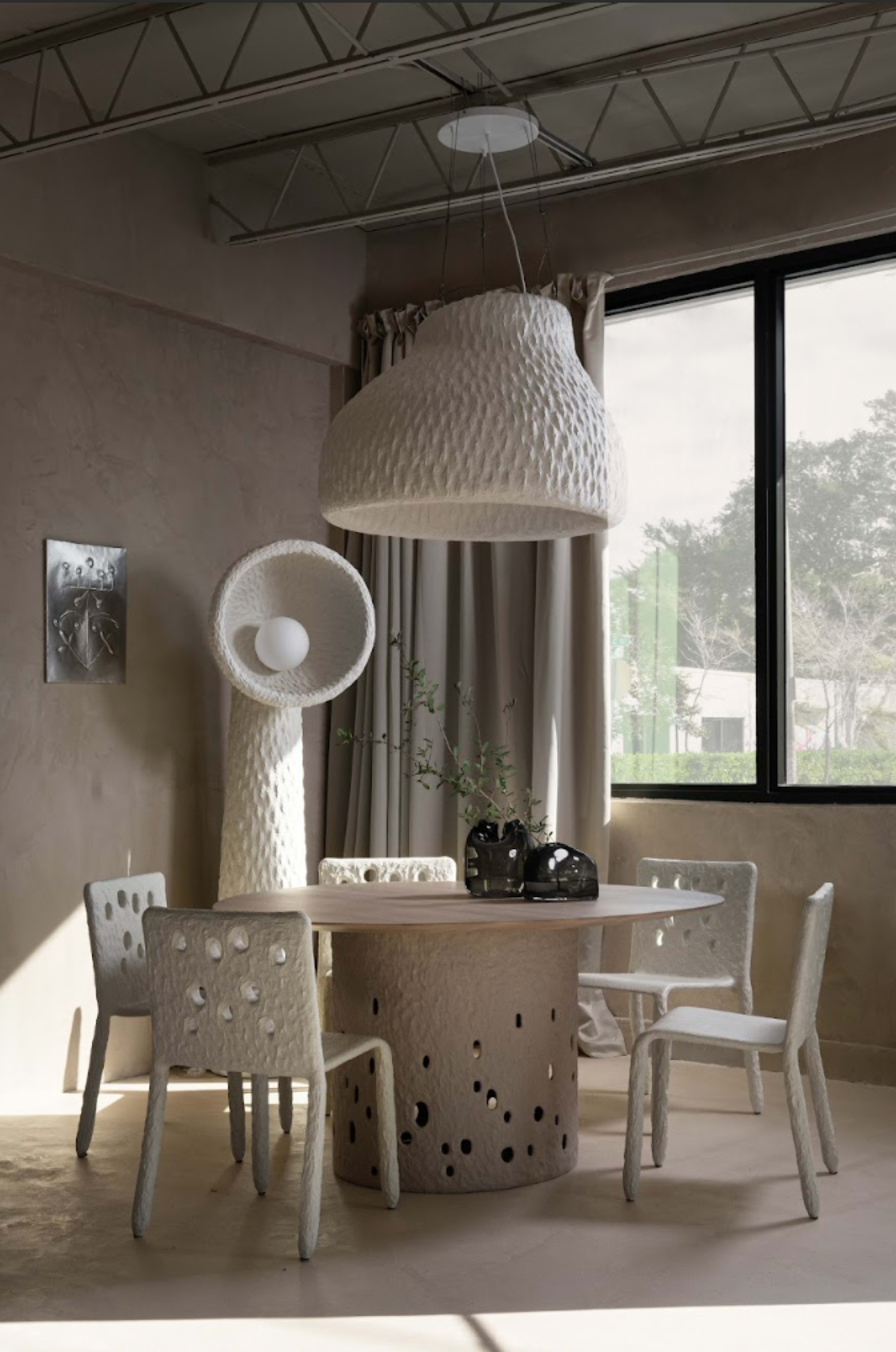 A modern dining area features a round table with a textured surface, surrounded by four unique chairs and illuminated by a sculptural pendant light.