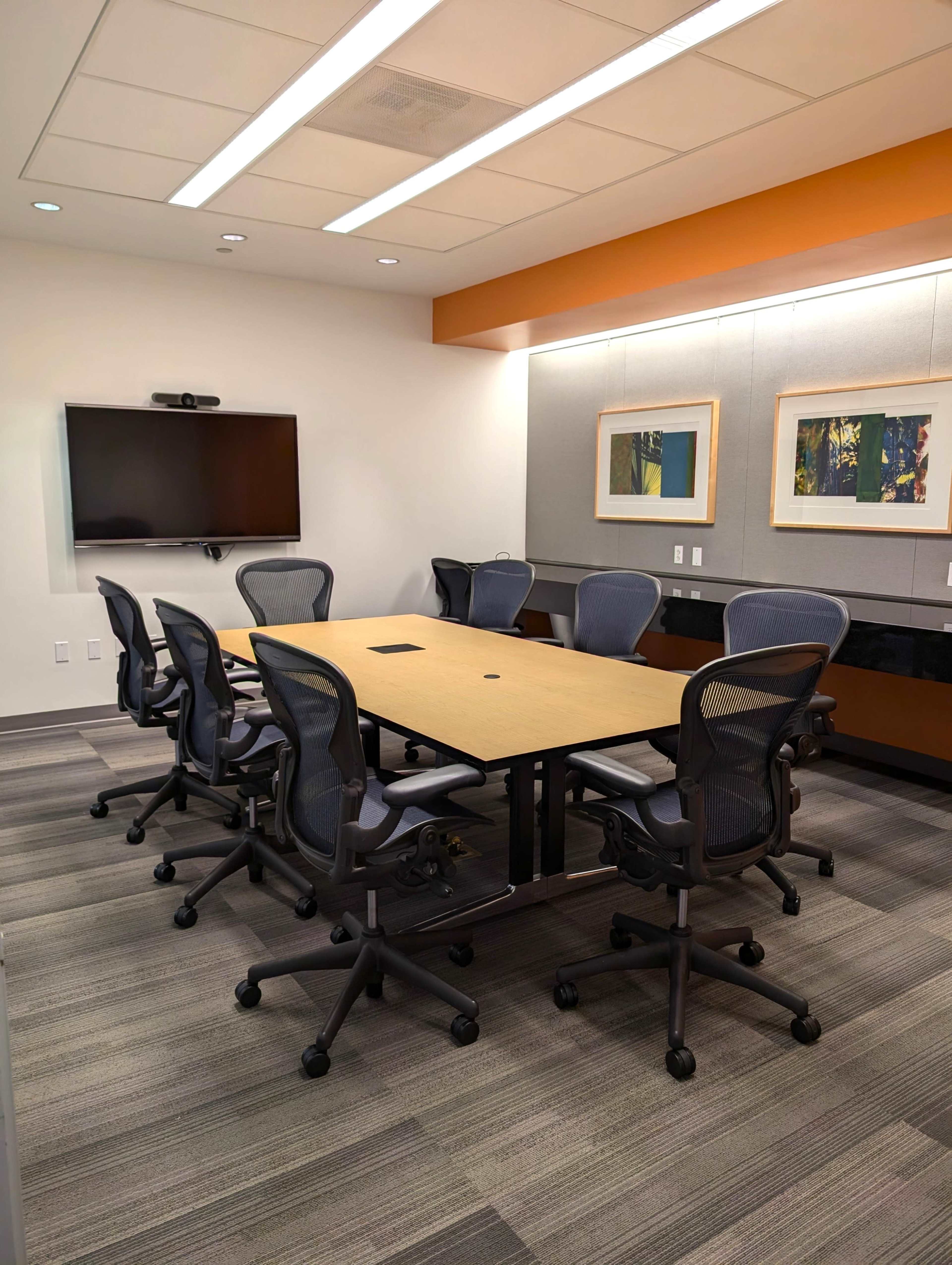 The image shows a modern conference room with a large table surrounded by several office chairs, a wall-mounted screen, and two framed artworks on the wall.
