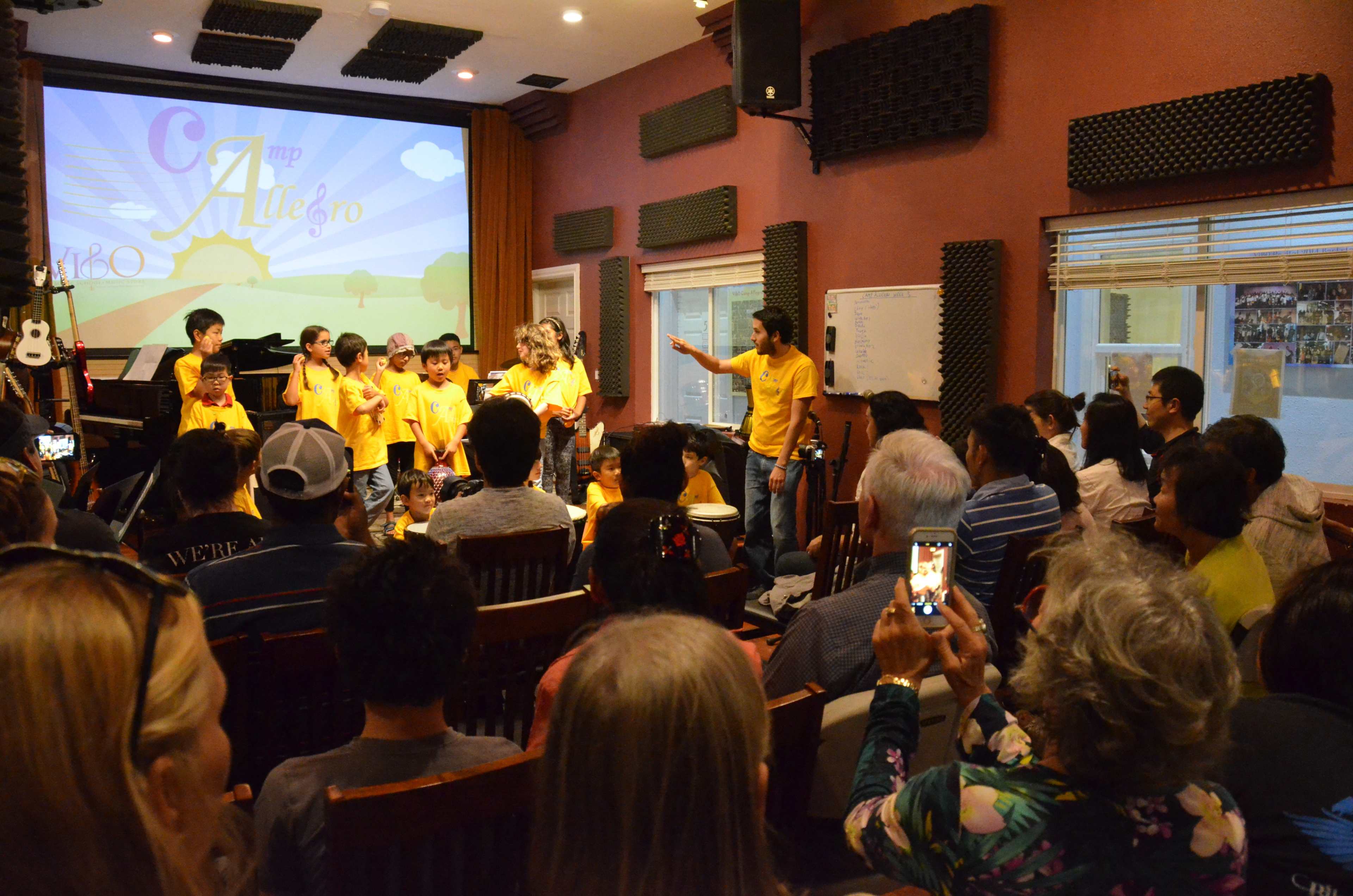 A group of children in yellow shirts performs on stage while an audience watches and records the event in a small auditorium.